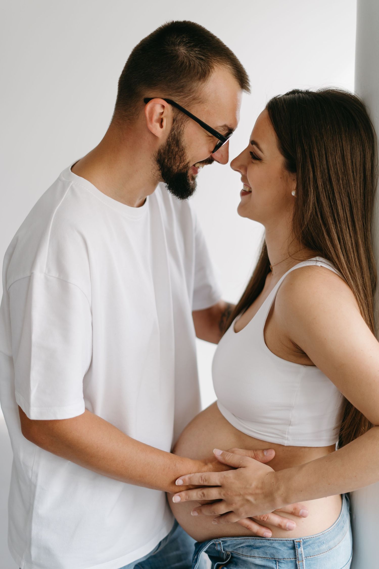 Intimate maternity photos of couple in white and denim showing tender moments against white wall.