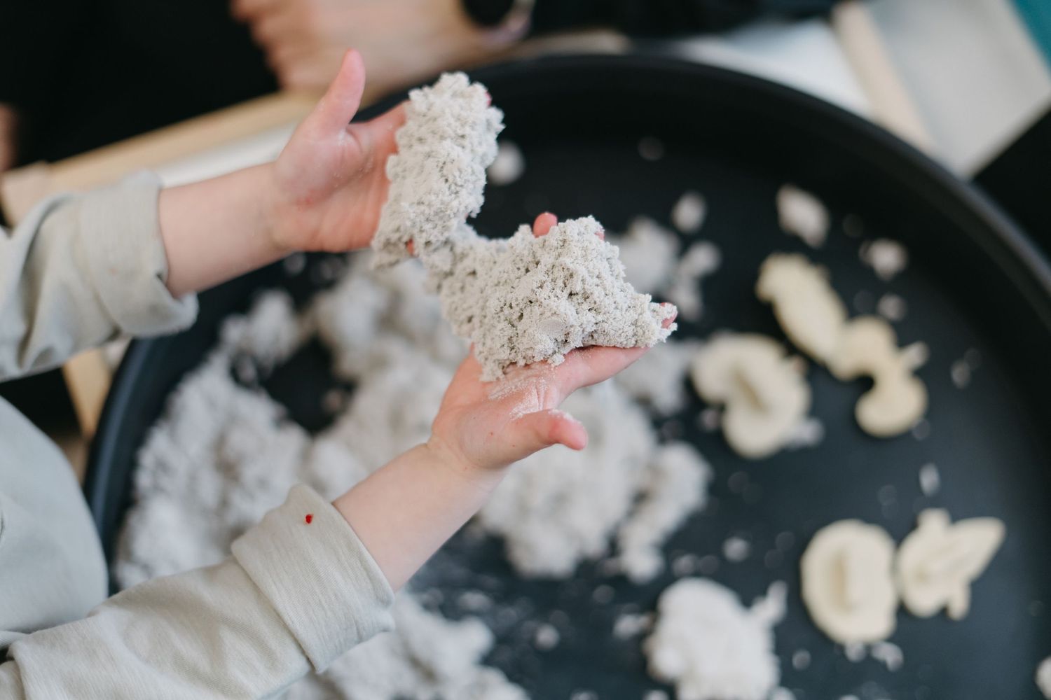 Hands holding clumps of raw sourdough dough above a dark pan with scattered flour during bread making process.