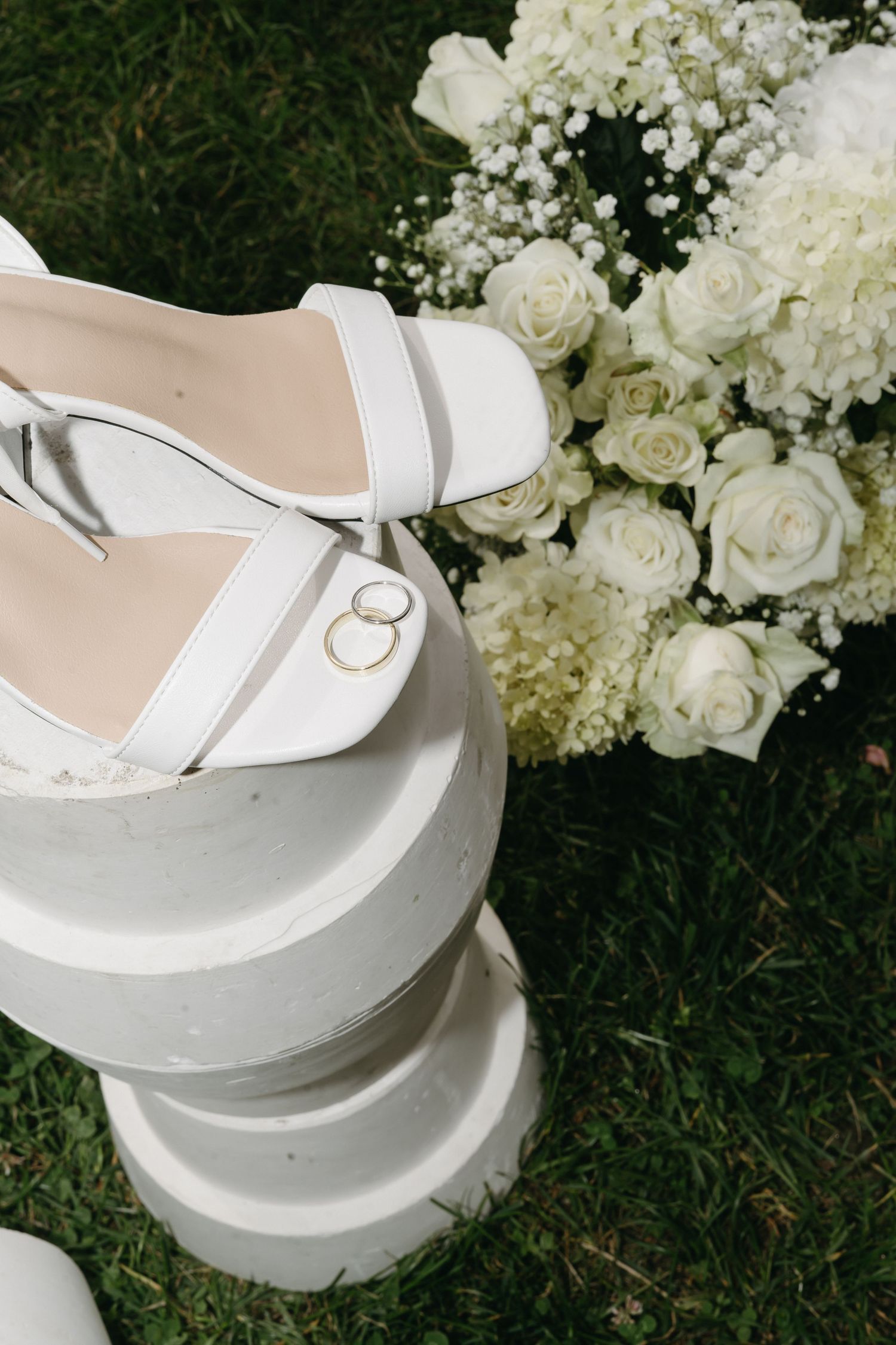 Wedding rings nestled among white roses and floral arrangements on a reflective surface, with bridal shoes in the background.
