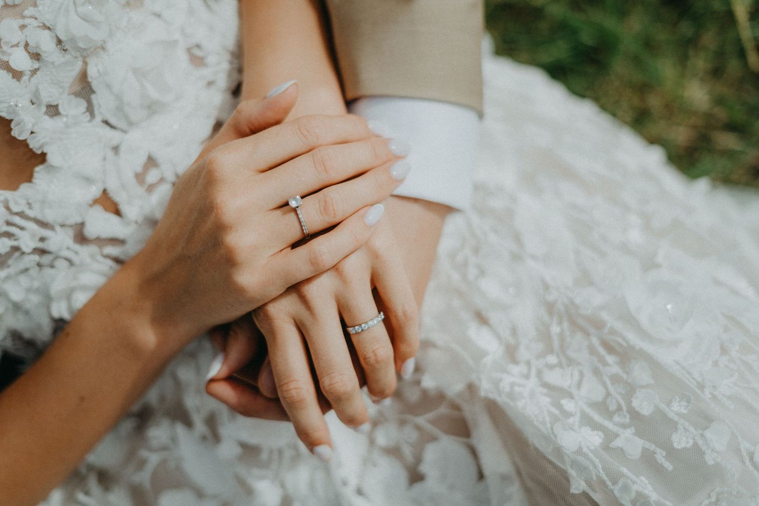 Close-up of hands with wedding rings resting on white lace dress showing romantic bridal details.