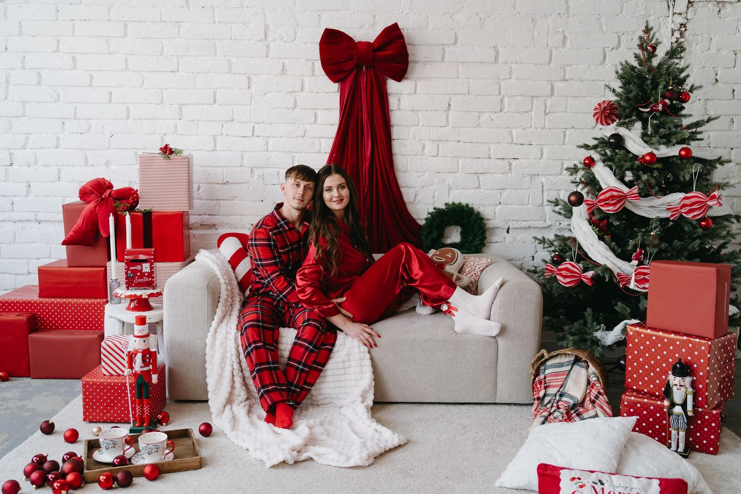 A couple in matching red plaid pajamas sits on a white sofa surrounded by Christmas gifts and a decorated tree.
