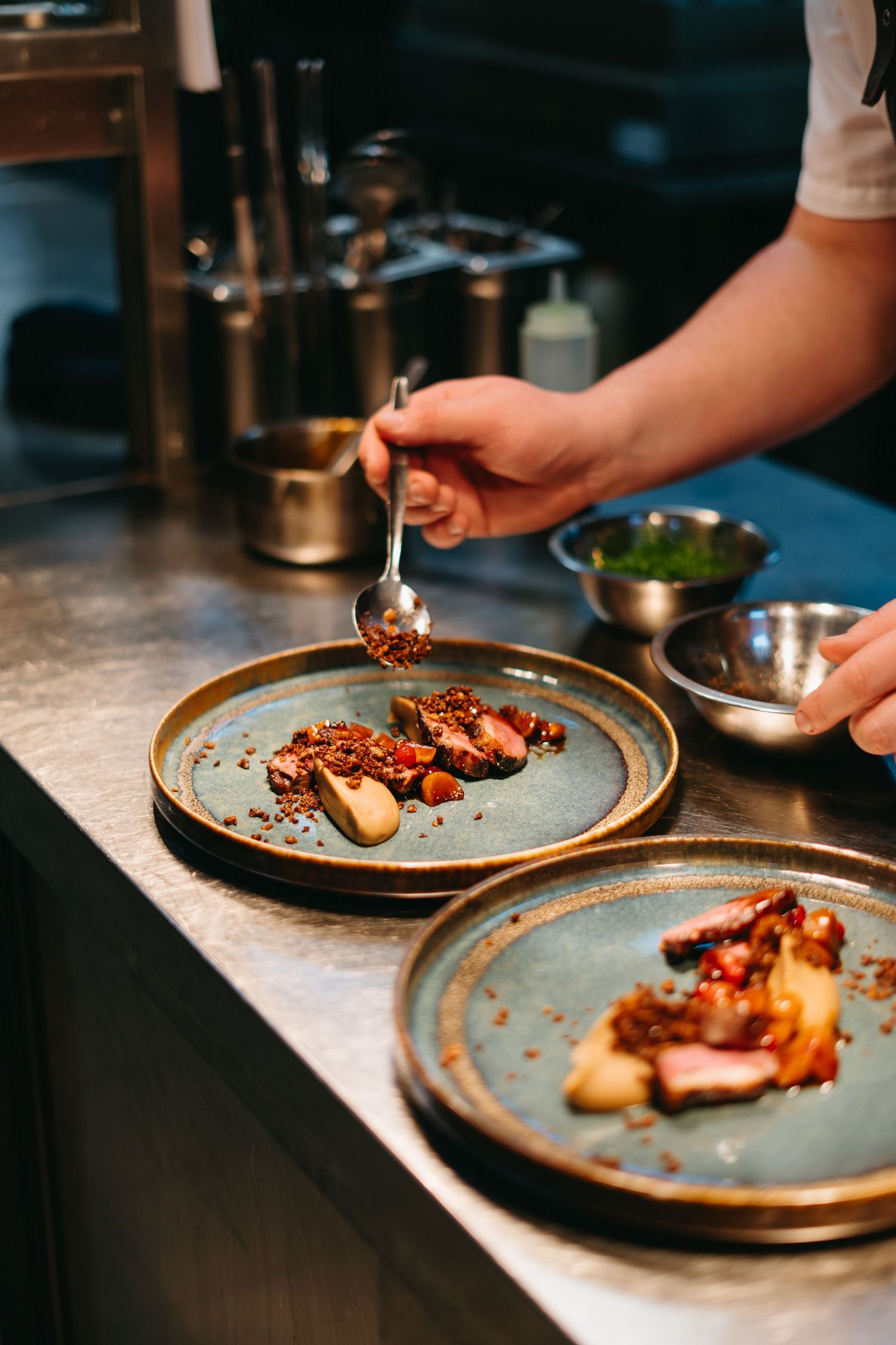 Chef plating an elegant dish with nuts and garnishes on blue ceramic plates at a restaurant kitchen counter.