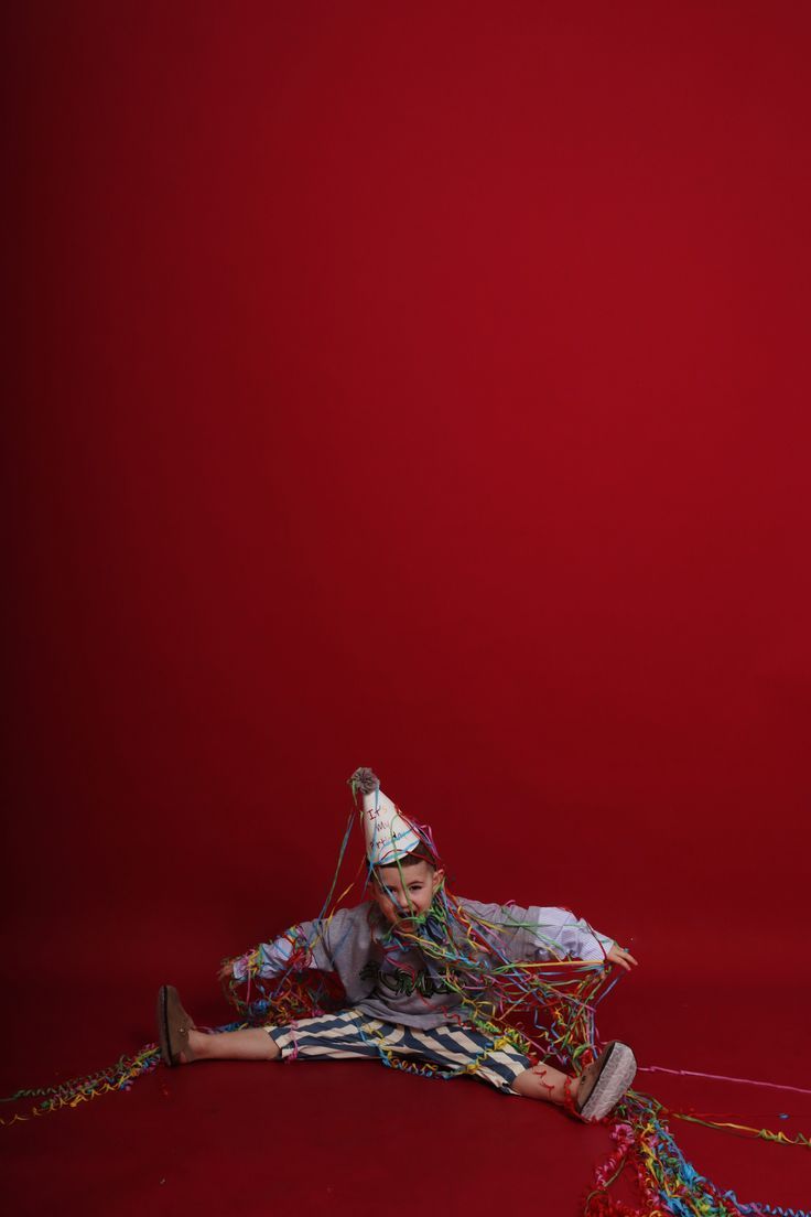A boy in a party hat sits on a red floor covered in colorful streamers and confetti.