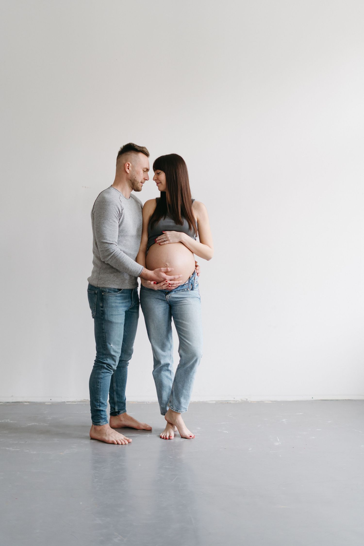Minimalist maternity photo showing a couple in casual denim standing close together against white wall.