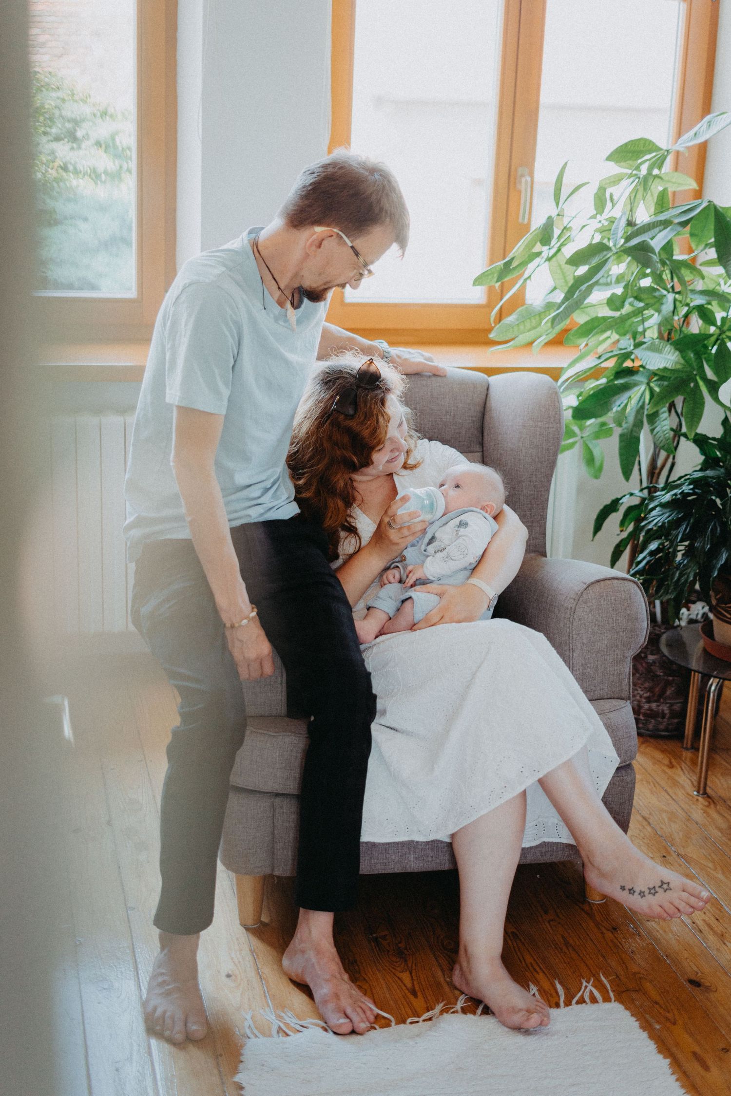 A couple admires their baby while sitting in a grey armchair near sunny windows and plants.