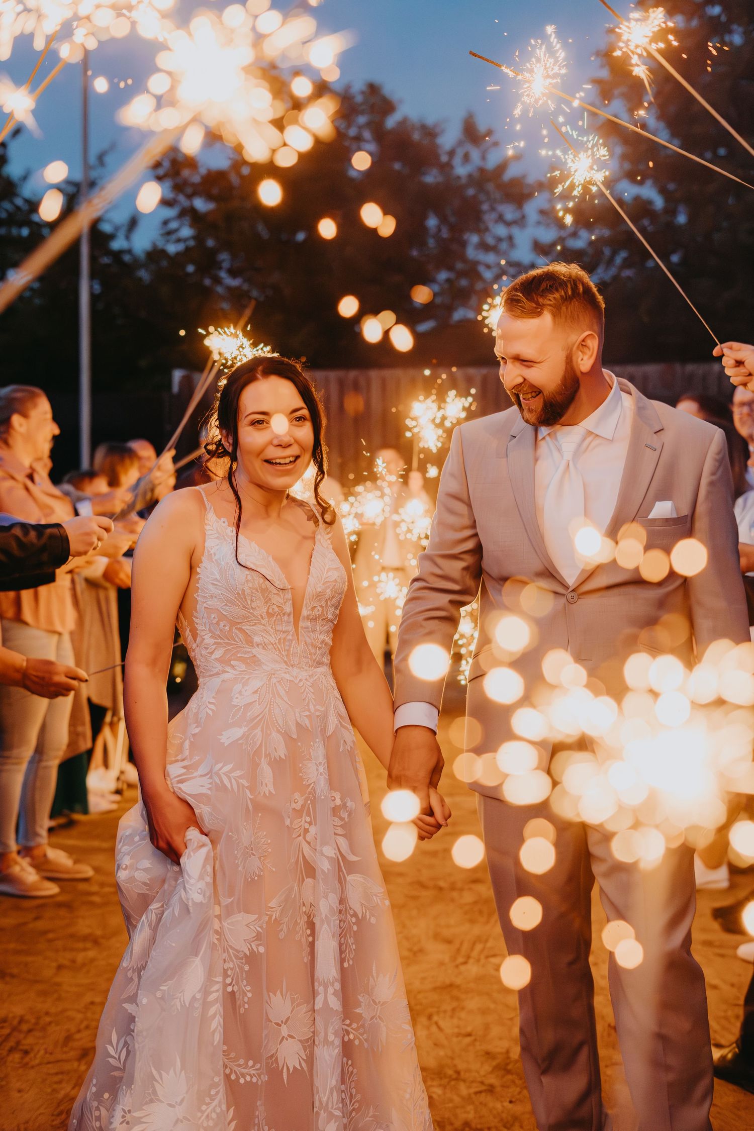Newlywed couple walks through sparkler exit at twilight wedding celebration with glowing sparklers creating magical atmosphere.