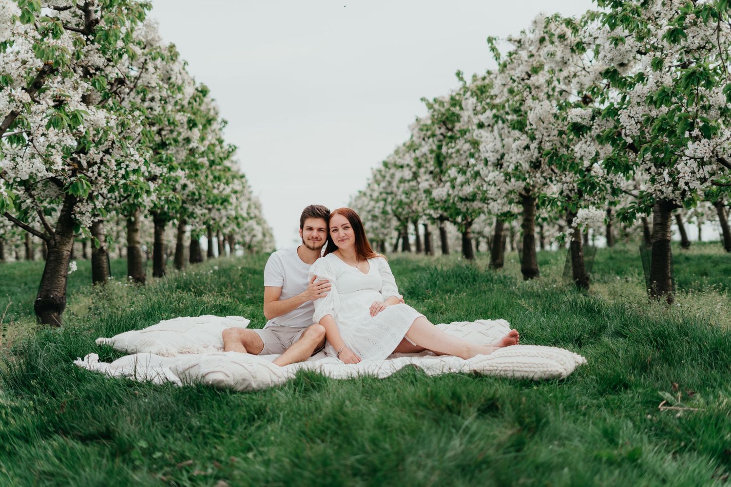 Couple sitting on white blanket in orchard surrounded by blooming spring trees.