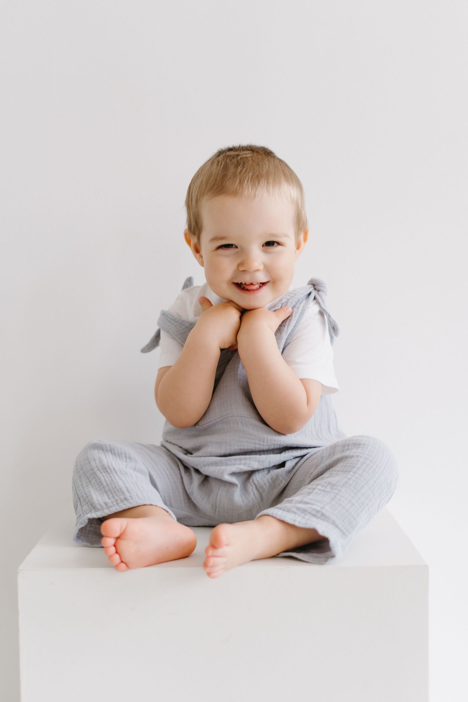 Toddler wearing gray outfit sits on white cube with joyful expression.