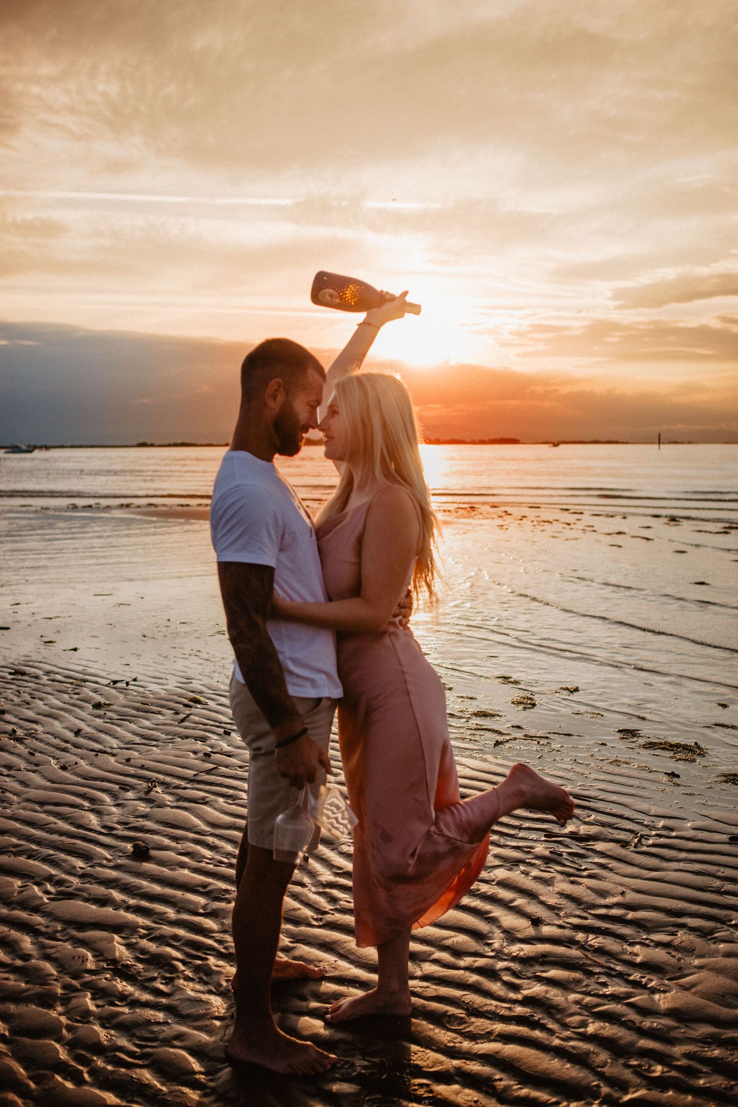 Romantic couple posing with champagne bottle on beach during golden hour.