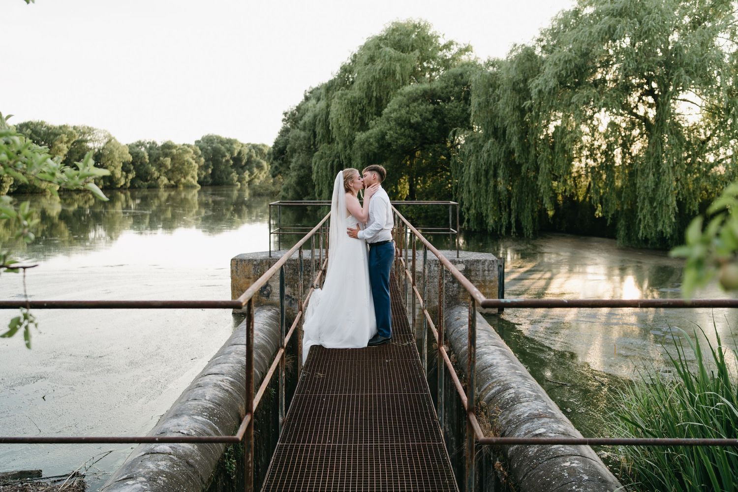 Bride and groom share a romantic kiss on a rustic dock overlooking a serene lake surrounded by willow trees.