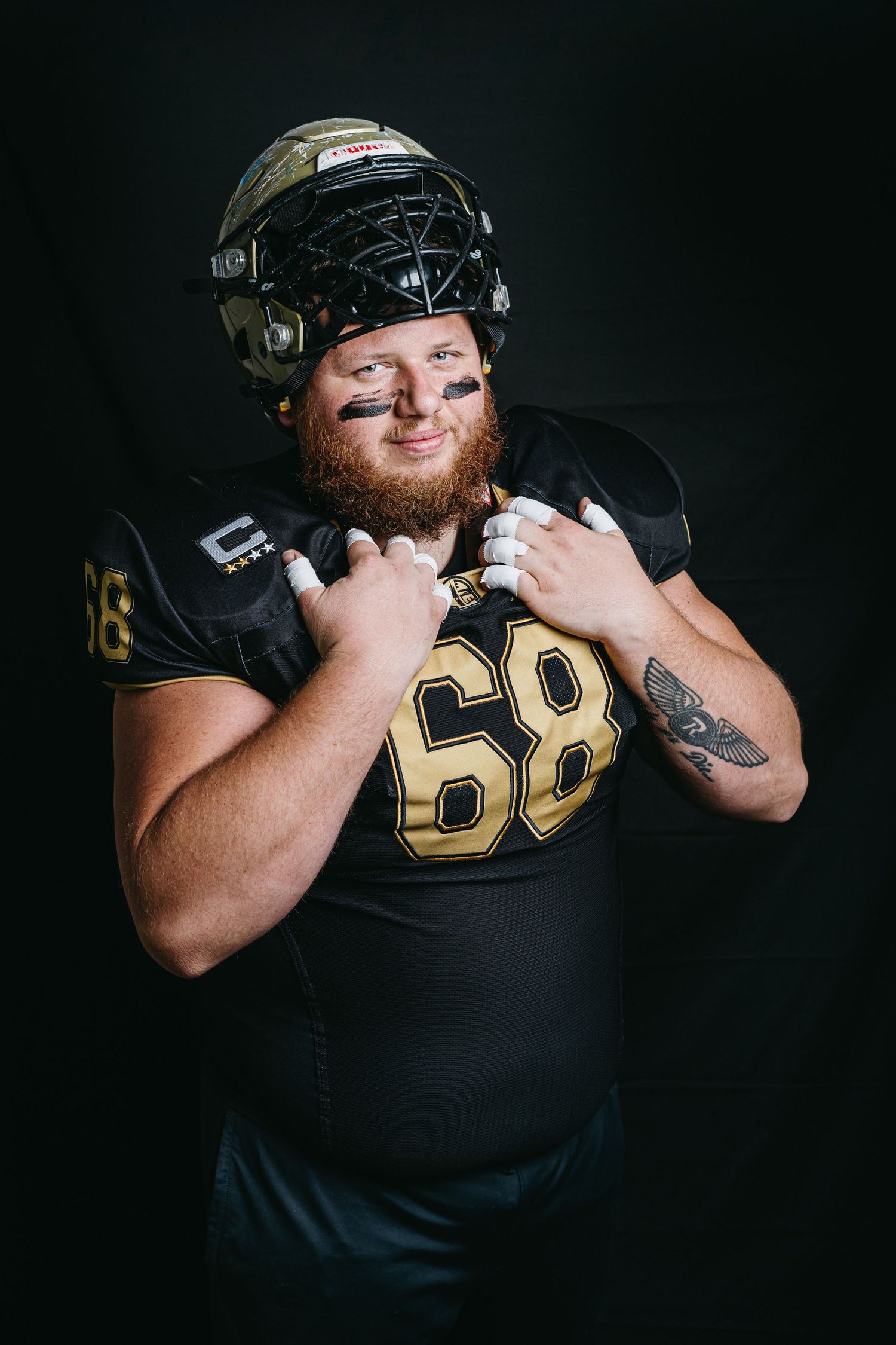 Football player in black uniform with gold number 68 poses against dark background holding jersey straps.