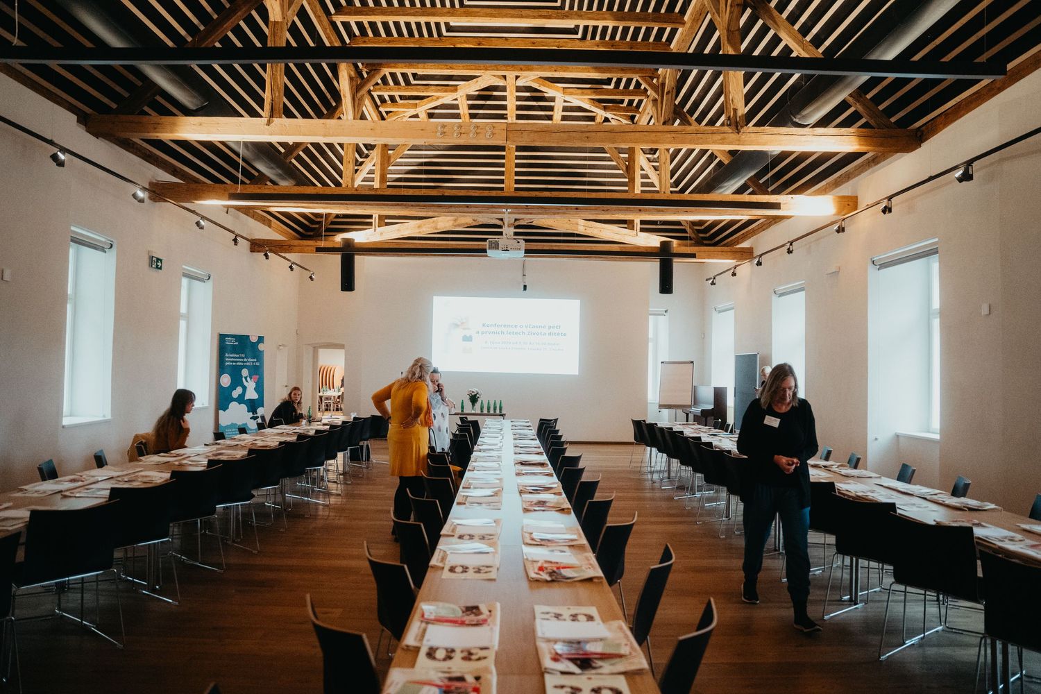 Long dining tables set up in modern venue with exposed wooden beams and windows for business conference or meeting.