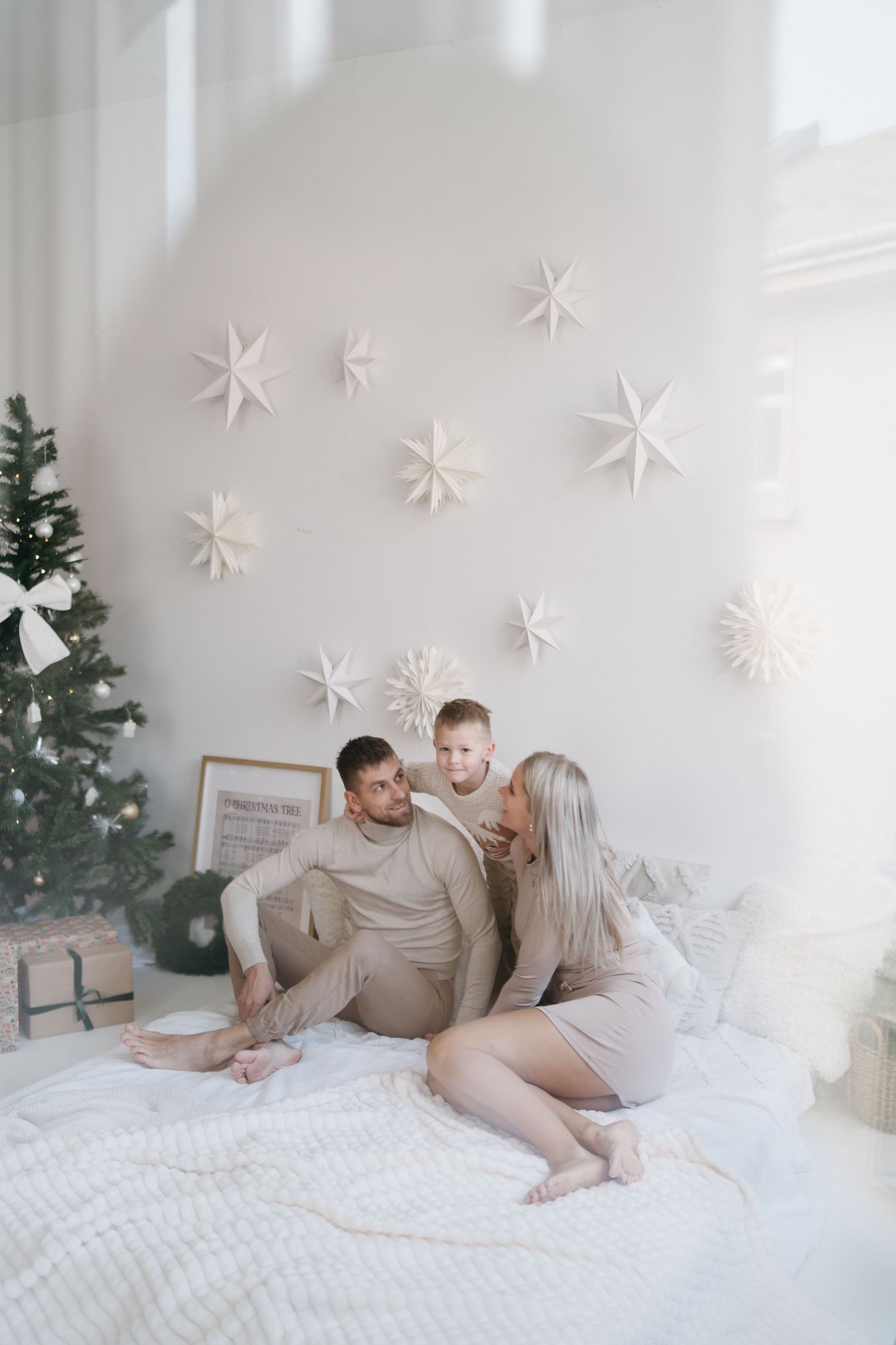 Family in beige outfits sitting on white bed with Christmas tree and paper star decorations on wall.