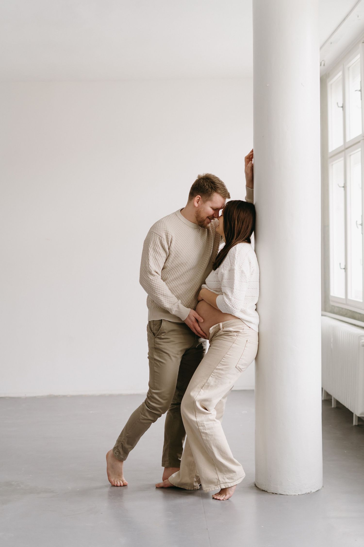 A romantic maternity moment captured against a white pillar in neutral-toned clothing.