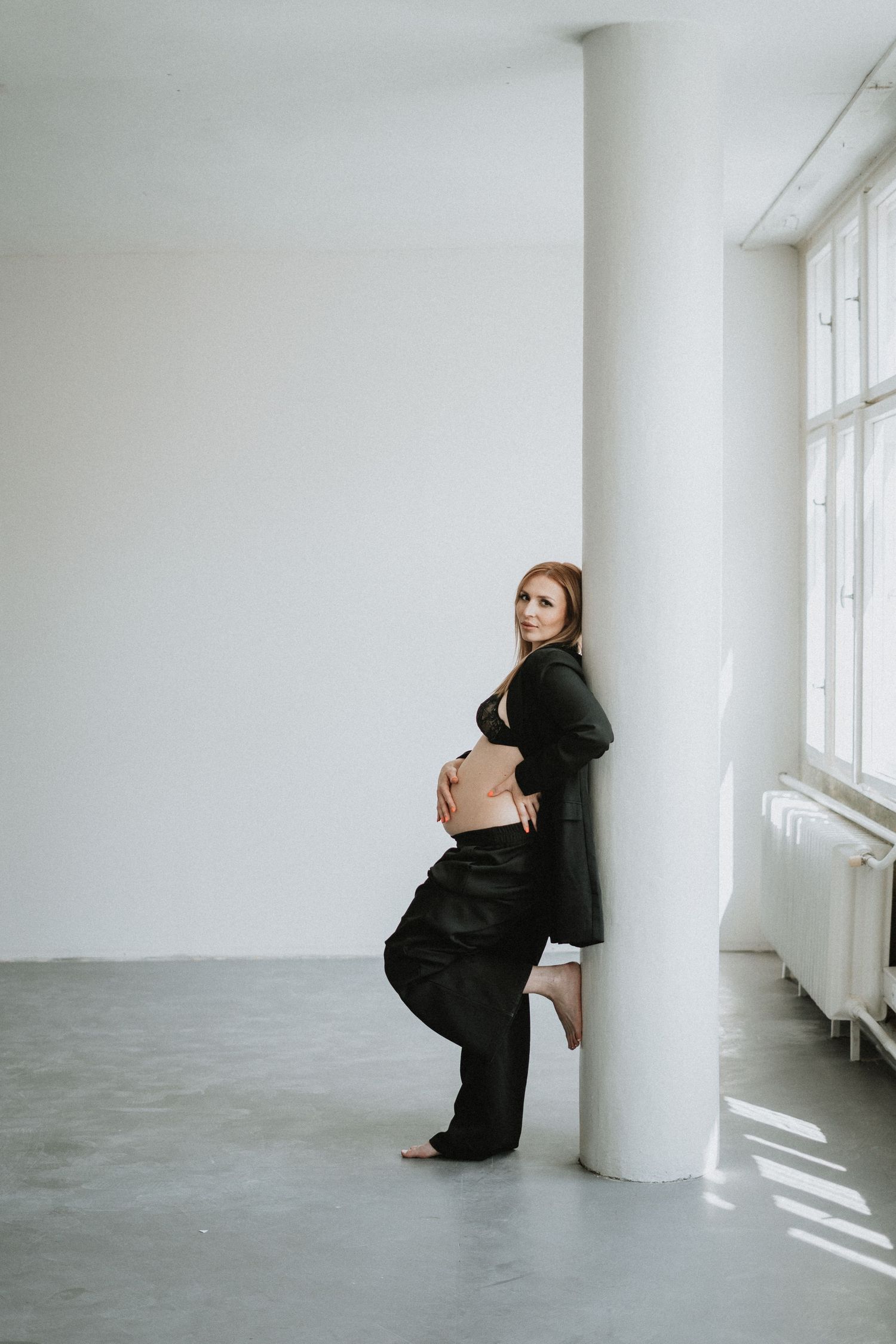 Artistic maternity photo of pregnant woman in black outfit leaning against white pillar.