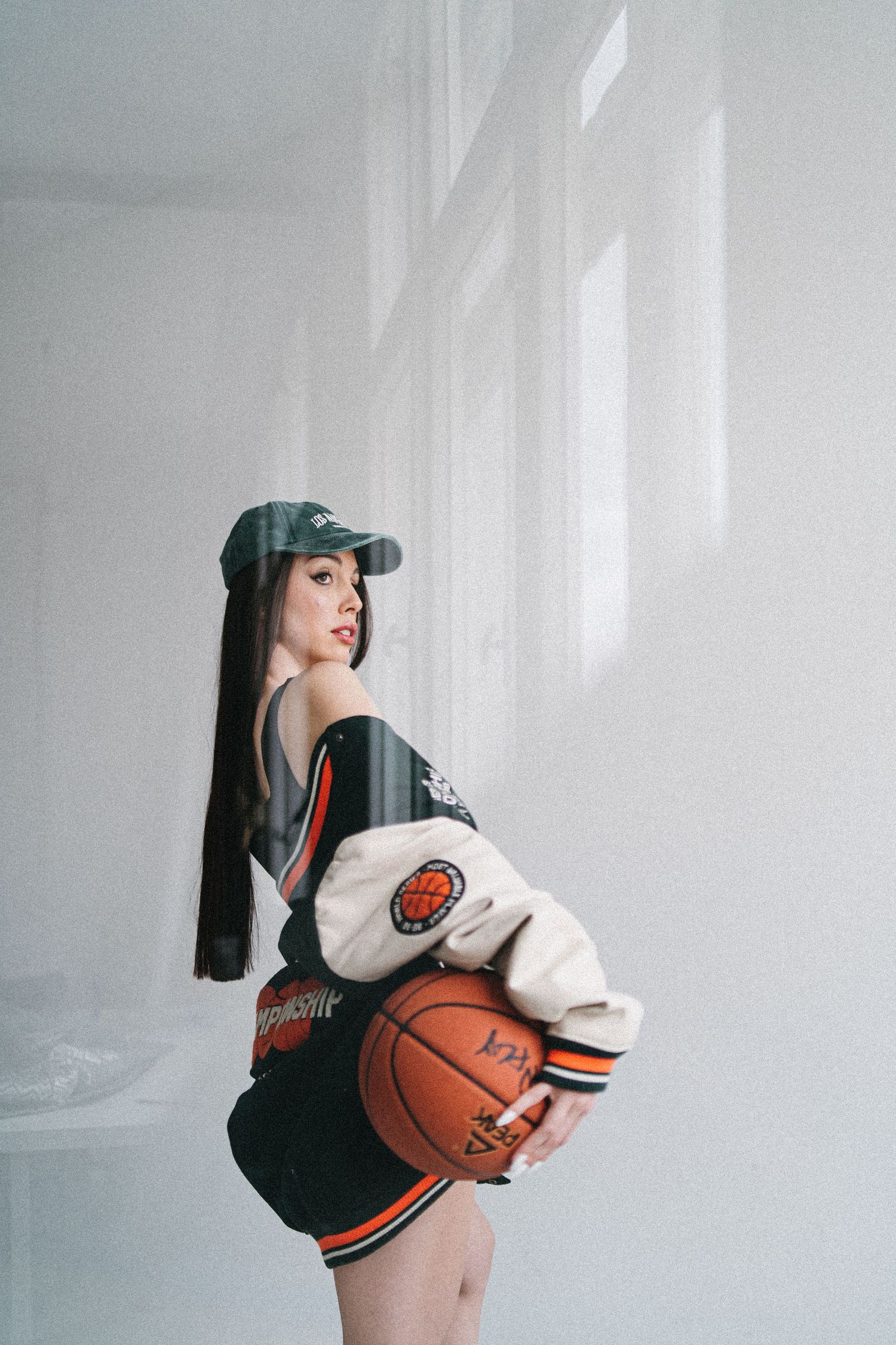 Young woman in streetwear holding a basketball, wearing a cap and varsity jacket in a bright white room.