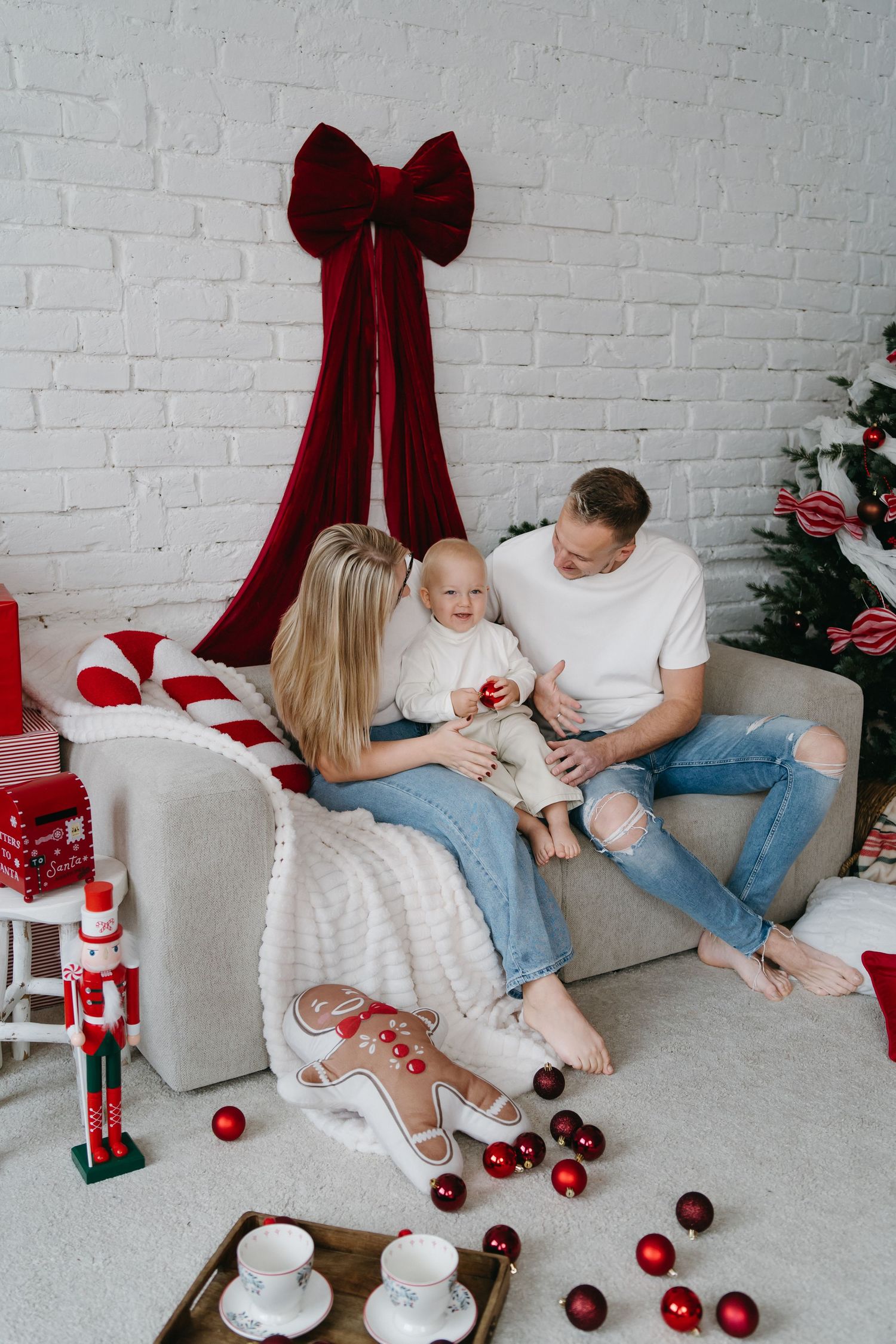 Happy family with baby celebrating Christmas on cozy sofa with red decorations, nutcracker, and festive tree.