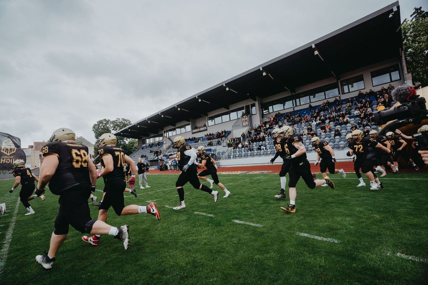 Rugby players in all black uniforms train on a grass field with spectator stands visible in the background.