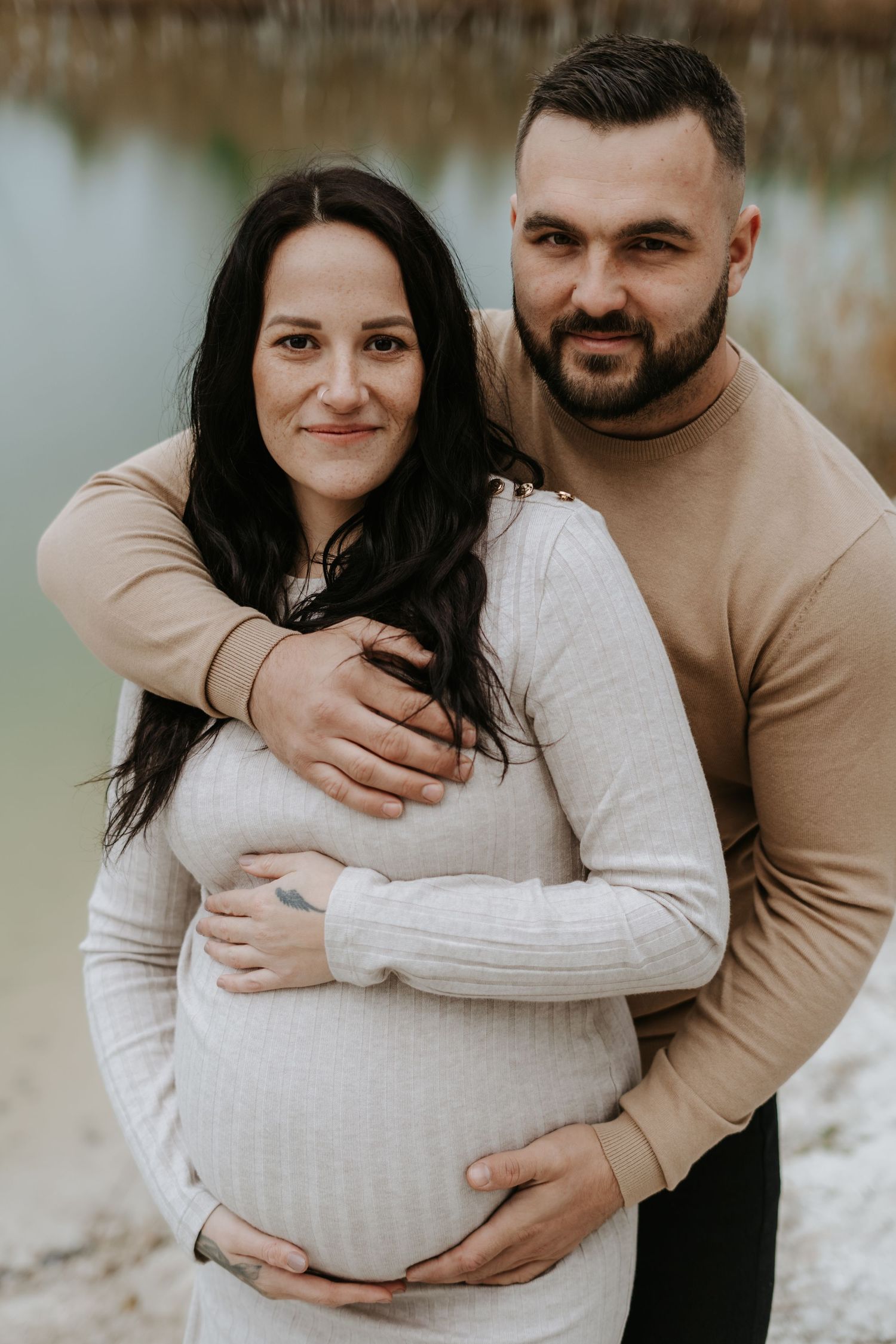 Couple in matching beige sweaters embrace during outdoor maternity session.