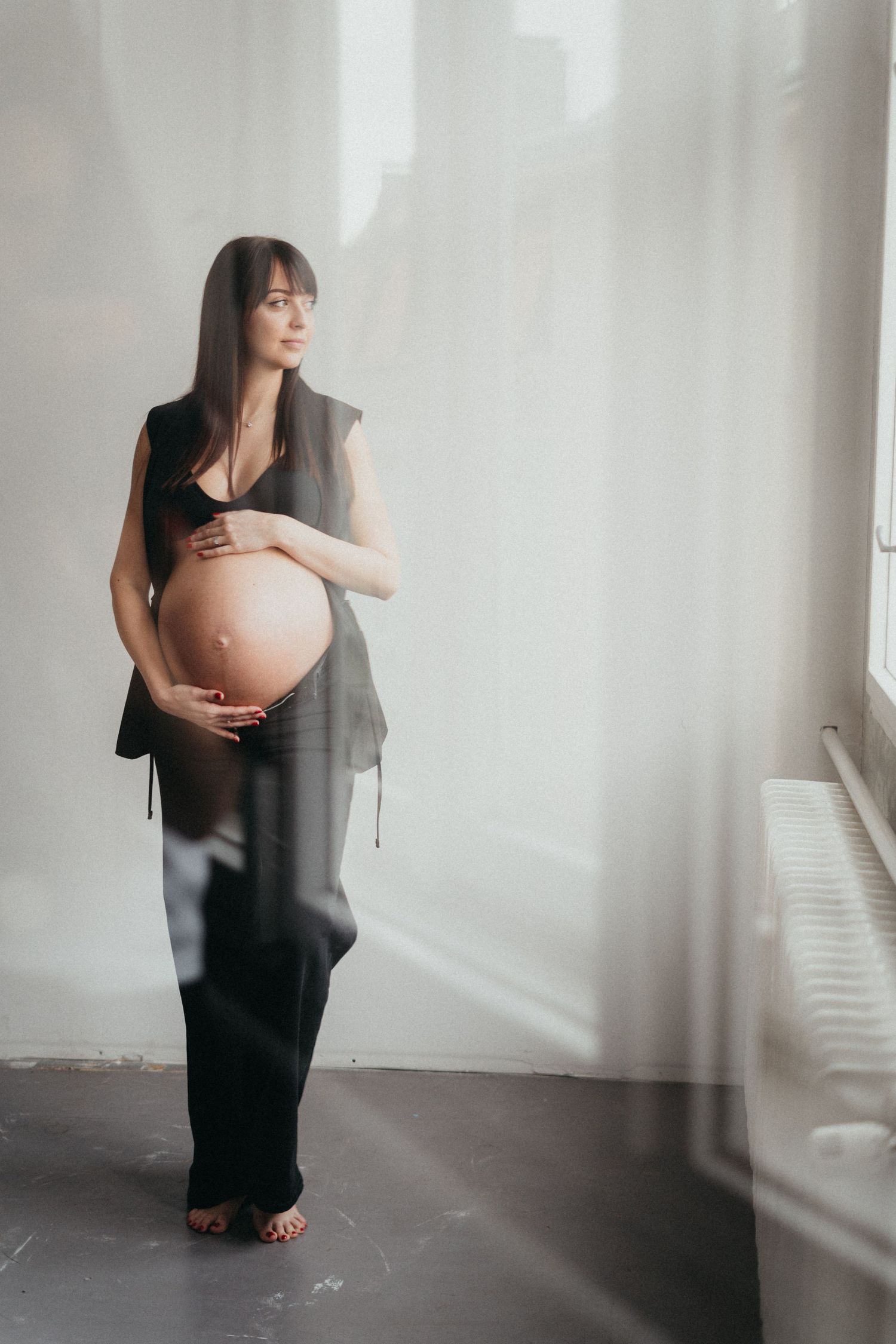 An expectant mother in dark clothing stands by a window, holding her pregnant belly in soft natural light.