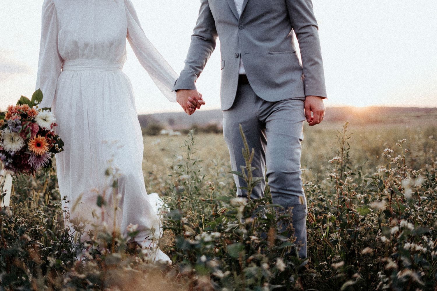 A couple holding hands walks through a wildflower field at sunset in wedding attire.