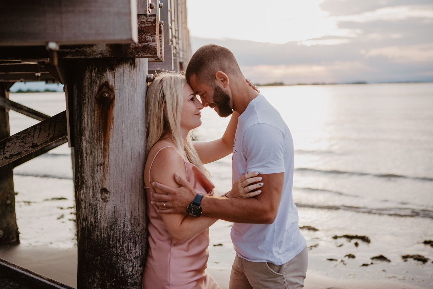 Romantic couple embraces near a wooden pier at sunset by the ocean.