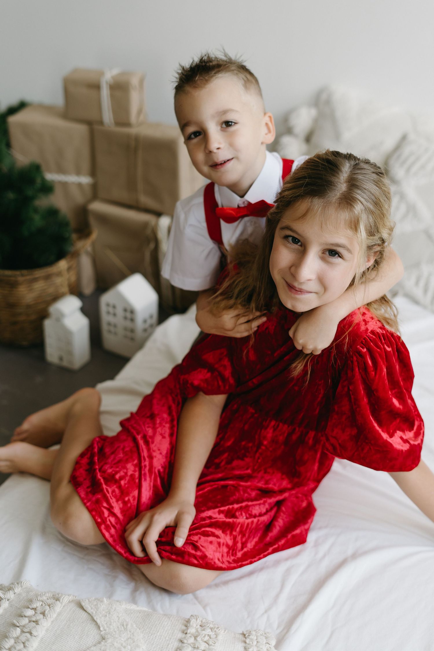 Children in red velvet and white outfits pose playfully on white bedding for a festive holiday photoshoot.
