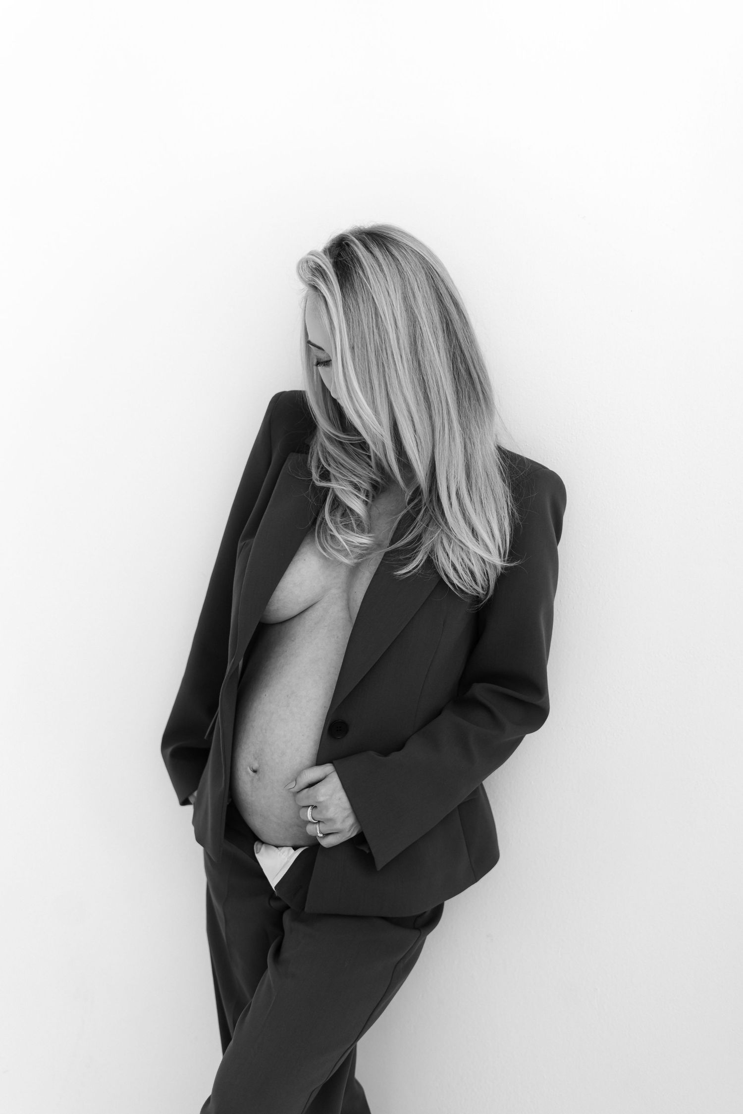 Black and white artistic portrait of a pregnant woman in a blazer suit posing against a white background.