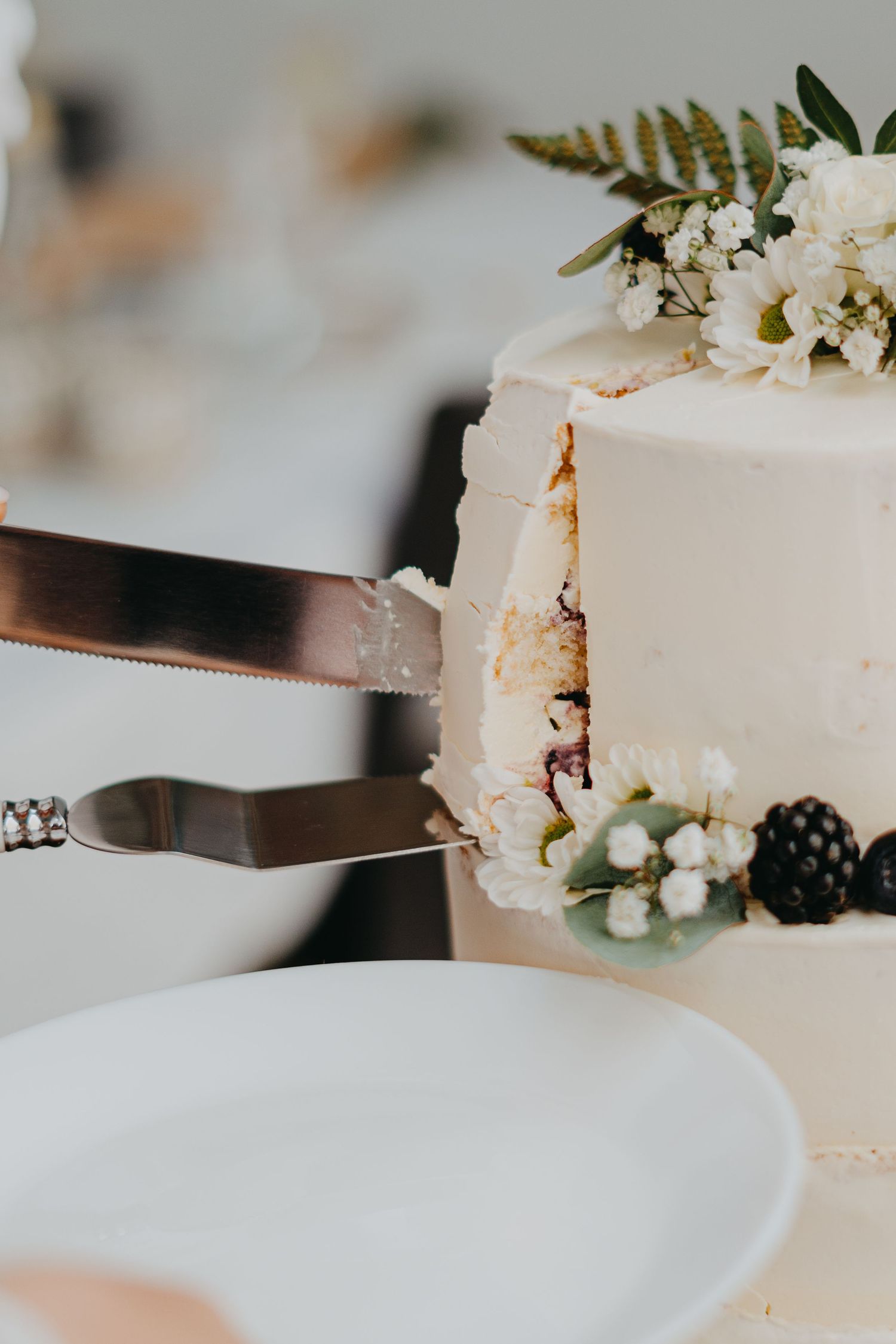 A silver knife cutting into a white wedding cake decorated with fresh flowers, blackberries and ferns.