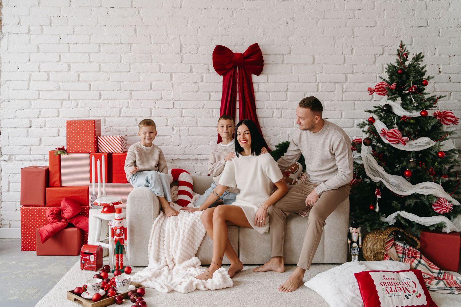 Family sits on white couch with Christmas tree, red presents and holiday decorations in modern minimalist setting.
