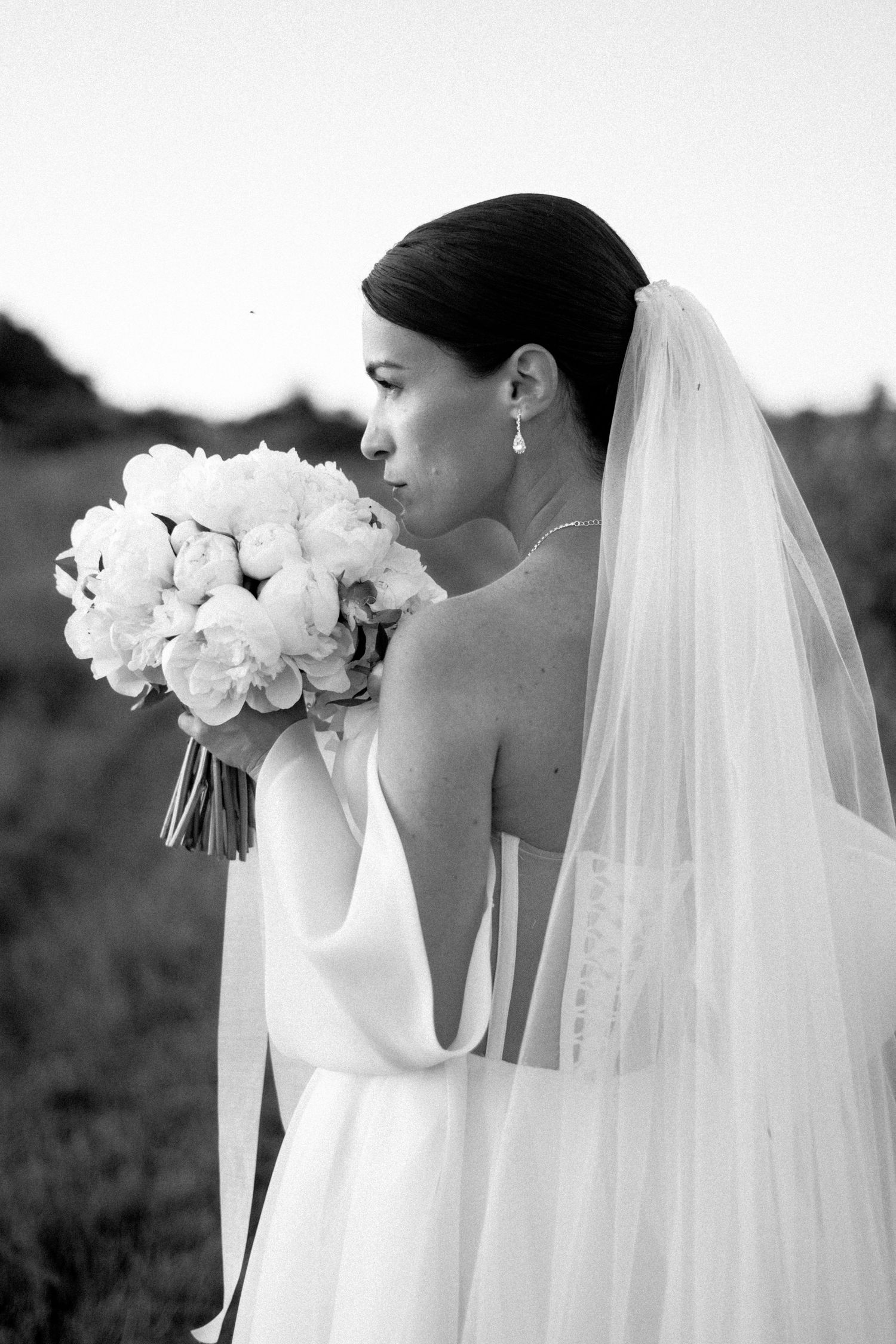 A black and white side profile of a bride wearing a long veil and holding a white bouquet of flowers.
