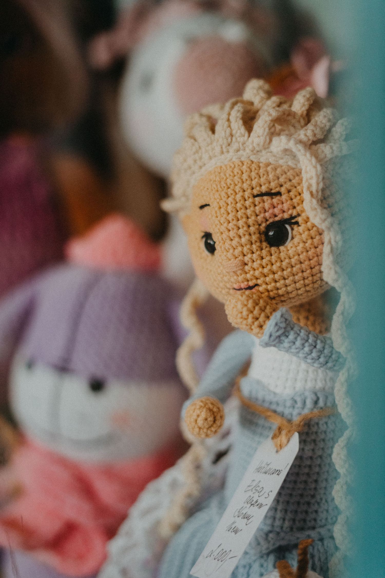 A handmade crocheted doll in a blue dress with blonde yarn hair shown in close up alongside other soft toys.
