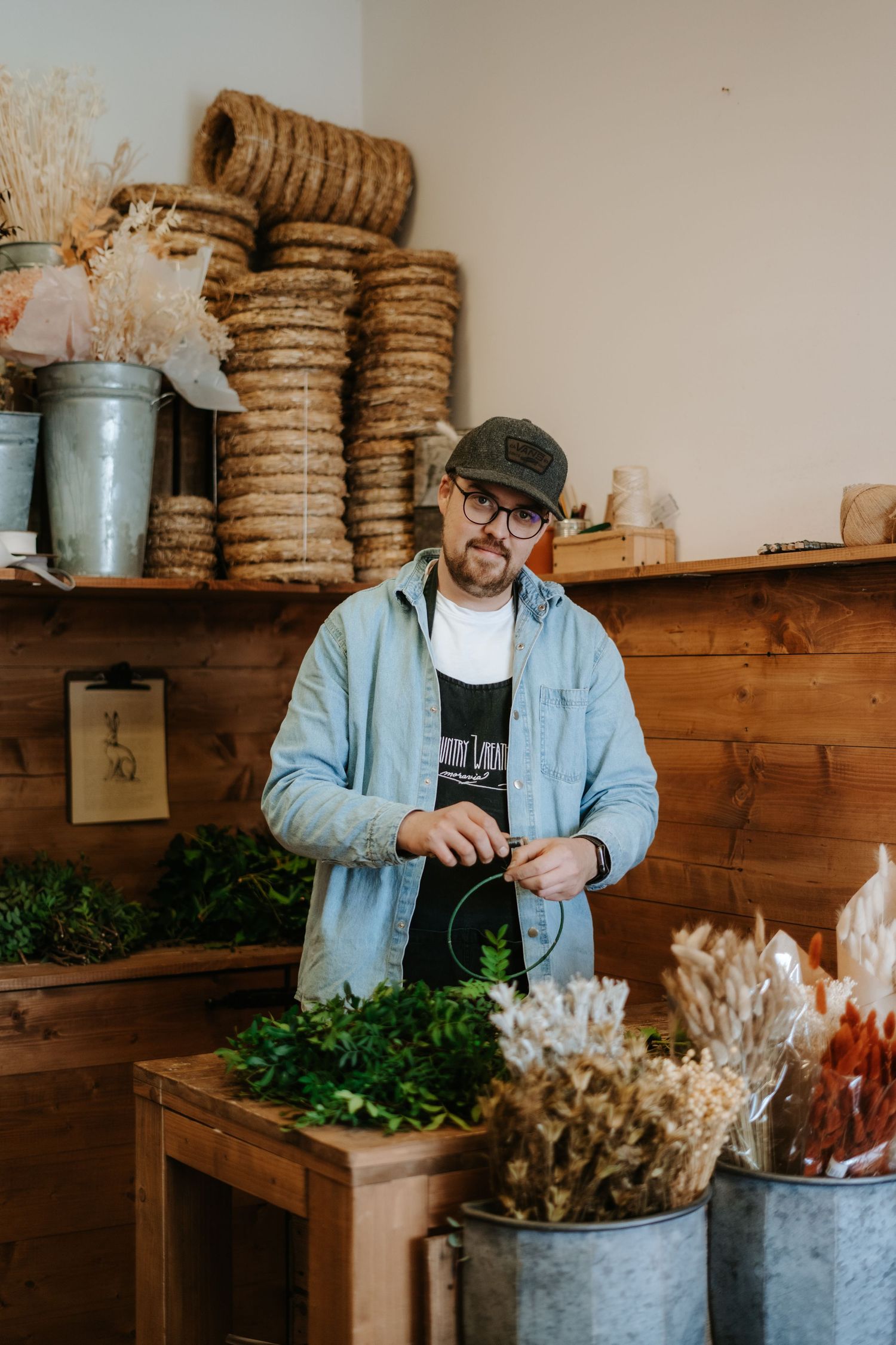 A shop worker arranges plants and dried flowers inside a rustic retail space with wooden shelves and vintage decor.