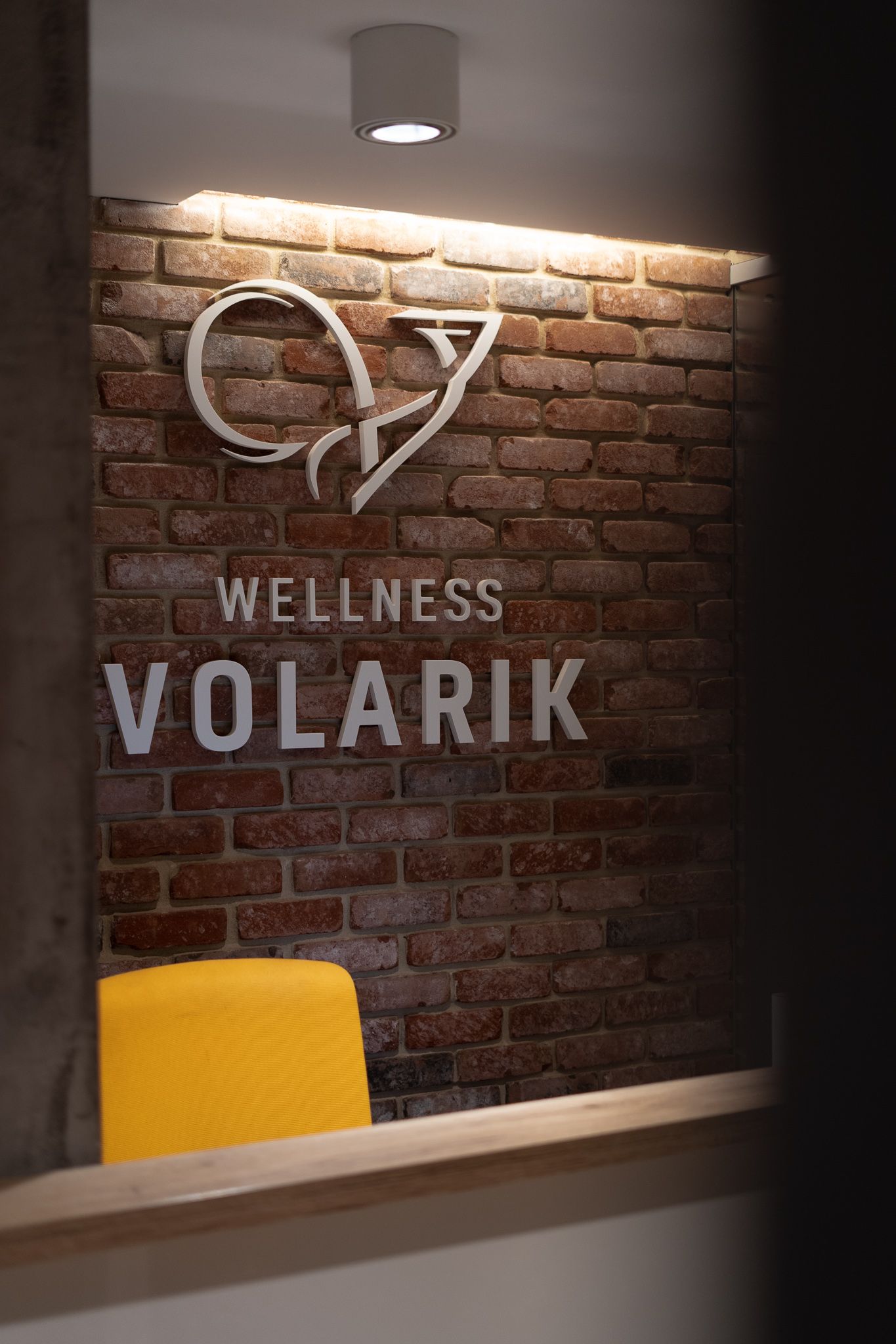 Illuminated signage for Wellness Volarik displayed on exposed brick wall with modern lighting and yellow accent decor.