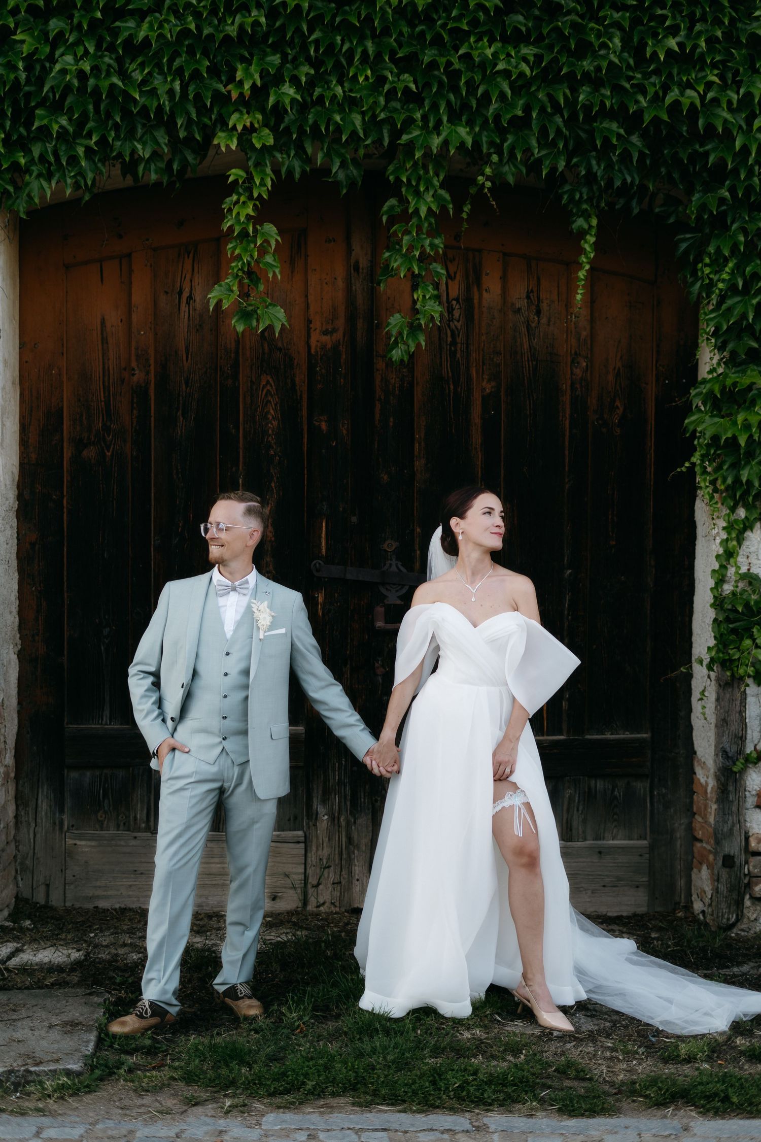 A couple in wedding attire poses back-to-back against a rustic wooden wall with green vines.