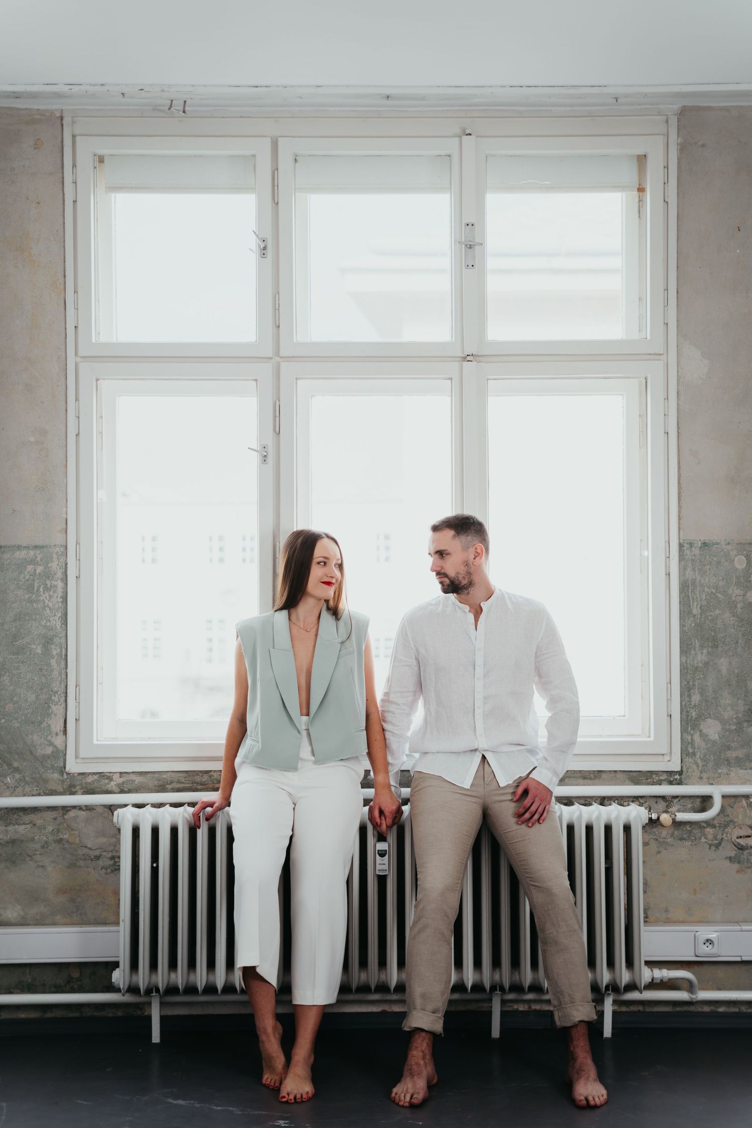 Barefoot couple in light clothing standing together by window and radiator.