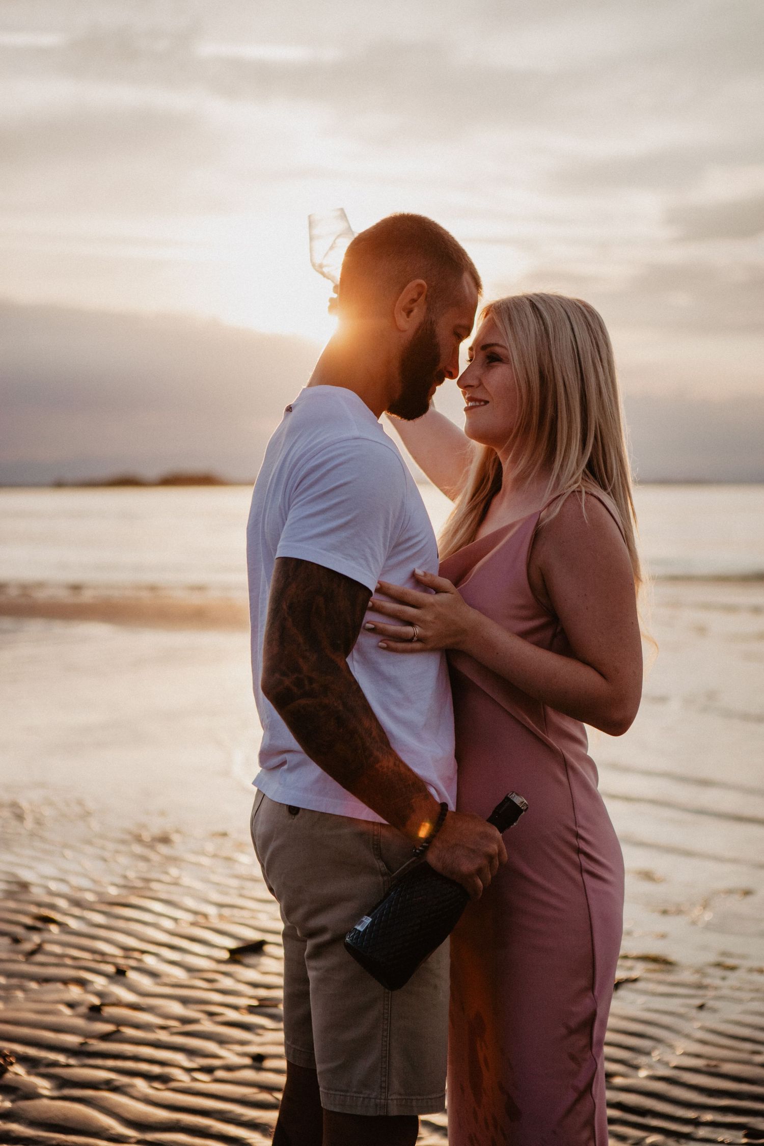 Intimate couple moment on beach at golden hour with person in pink dress.