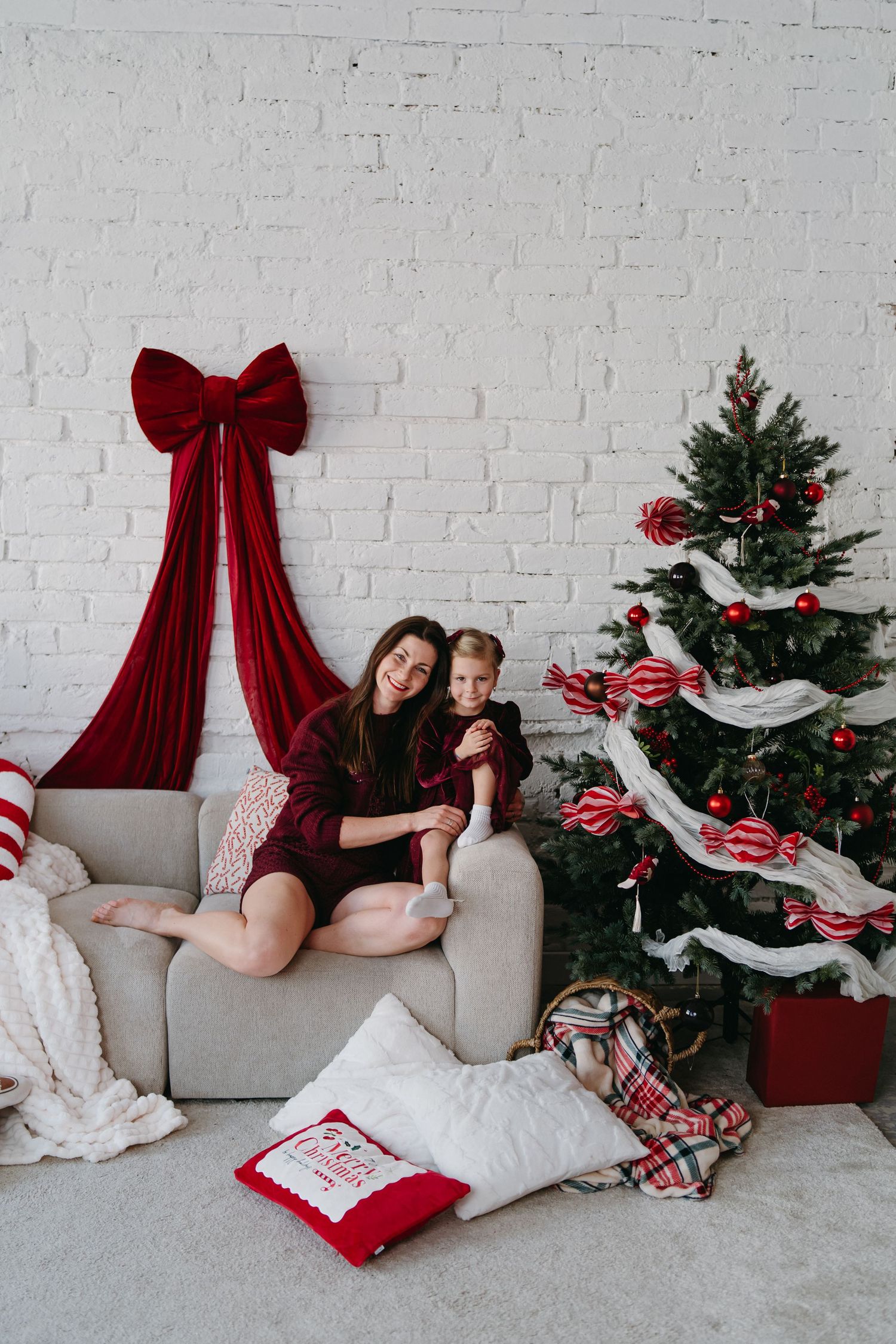 Mother and child in burgundy pajamas sitting on sofa beside decorated Christmas tree with red ornaments and large bow.