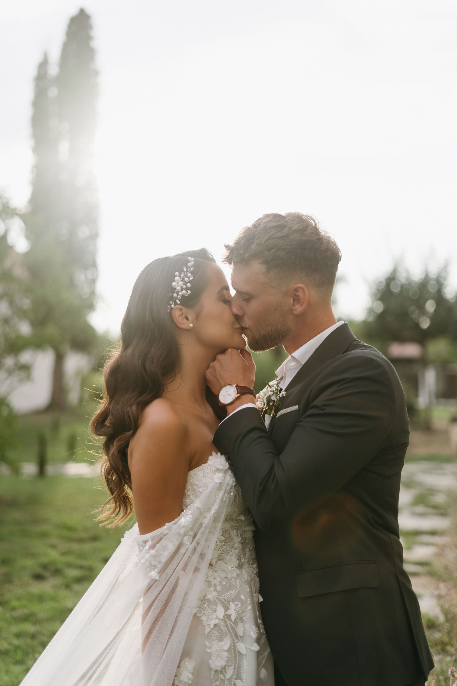 A couple shares a romantic kiss outdoors with sunlight streaming through trees in the background.