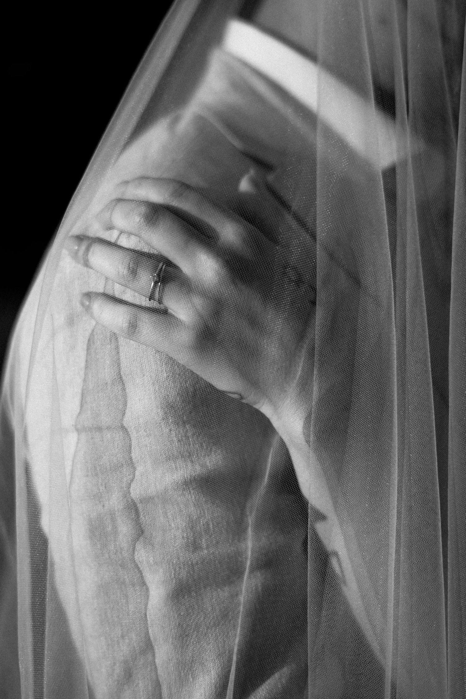 A black and white artistic photograph shows a hand wearing a wedding ring through delicate white veil fabric.