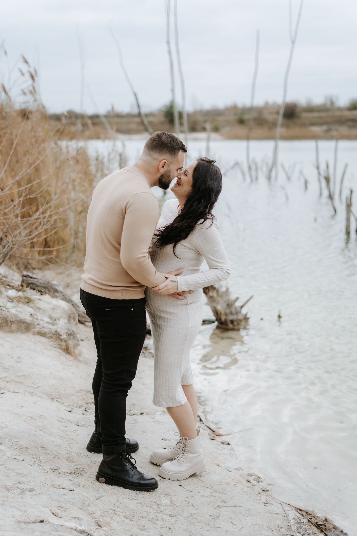Intimate maternity photoshoot by the water with couple in coordinating neutral outfits.