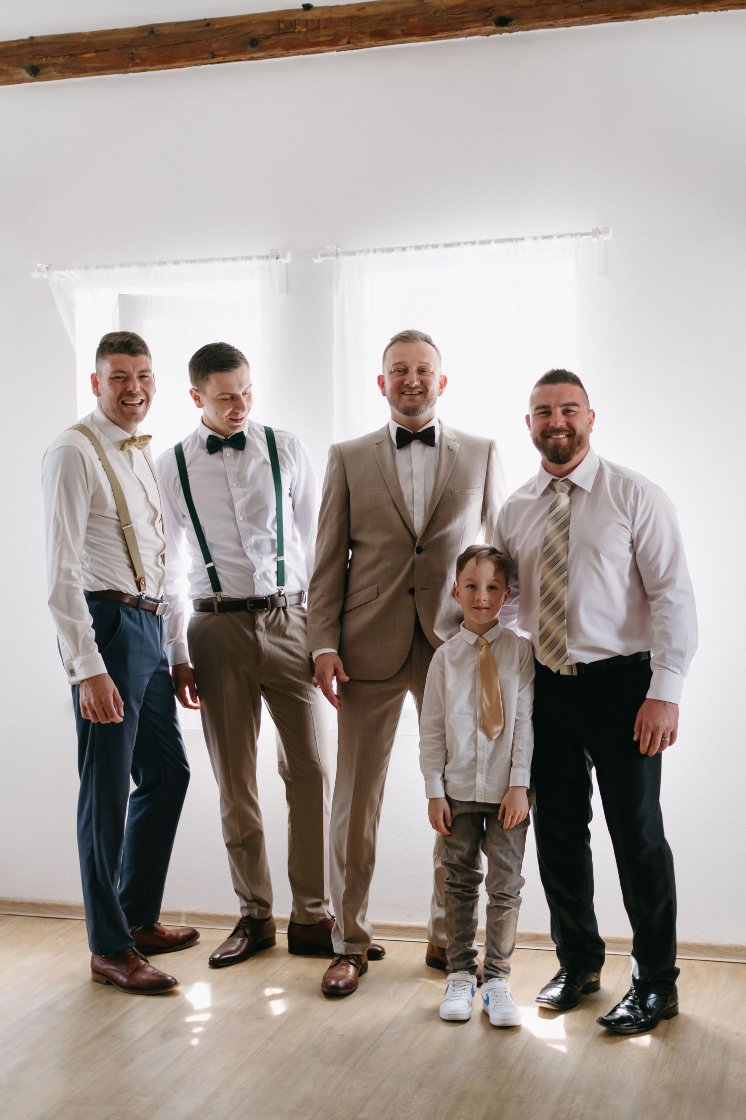 Five attendees dressed in formal attire with bowties, suspenders and suits pose together for a wedding party portrait.