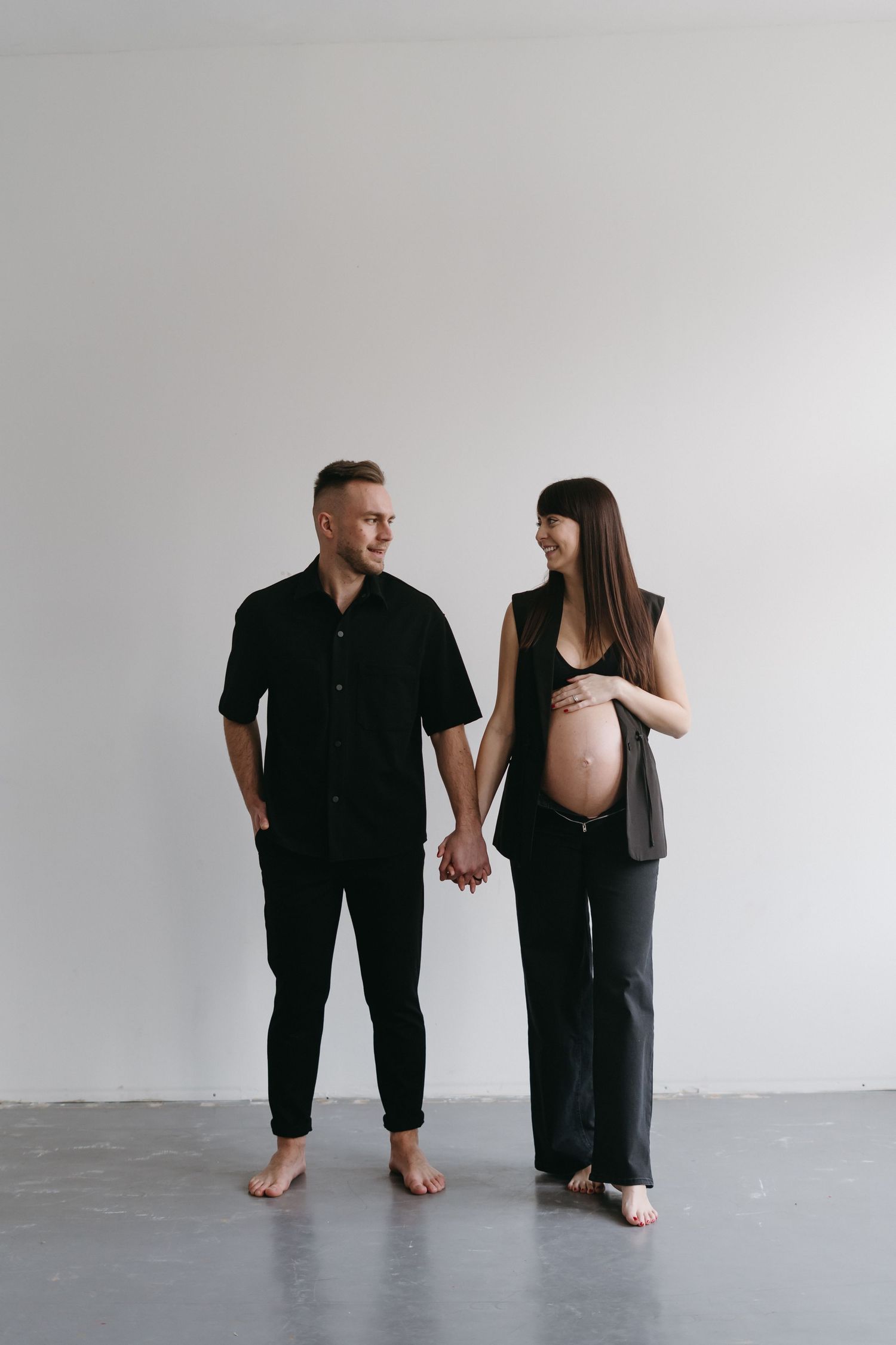 Minimalist maternity portrait of couple in black outfits holding hands against white wall.