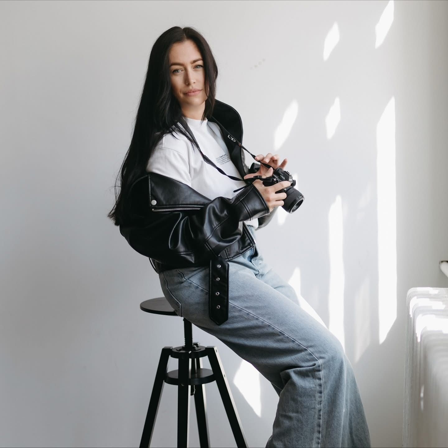 Woman with long dark hair in a black leather jacket and wide-leg jeans sits on a black stool holding a camera in a bright, minimalist room.