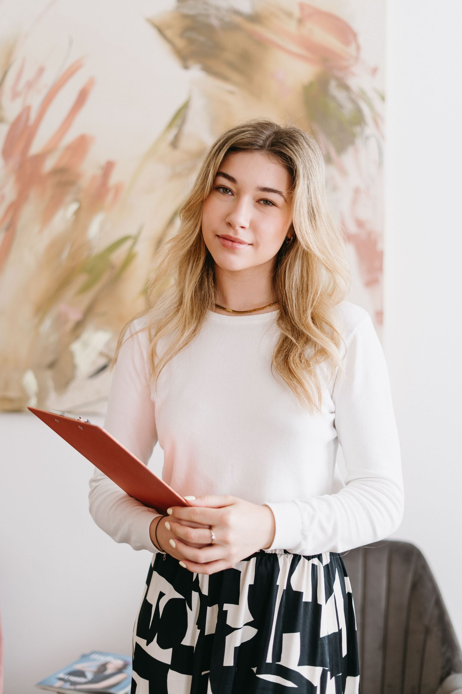Professional portrait against soft watercolor backdrop wearing white sweater and patterned skirt while holding red folder.
