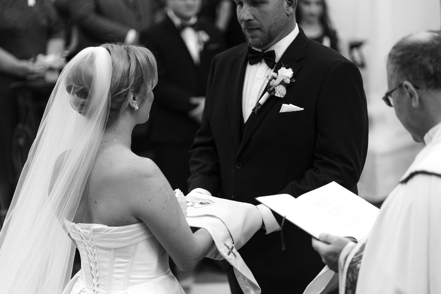Black and white photo of a bride in a strapless corset gown and veil facing a groom in a tuxedo during a wedding ceremony with an officiant.