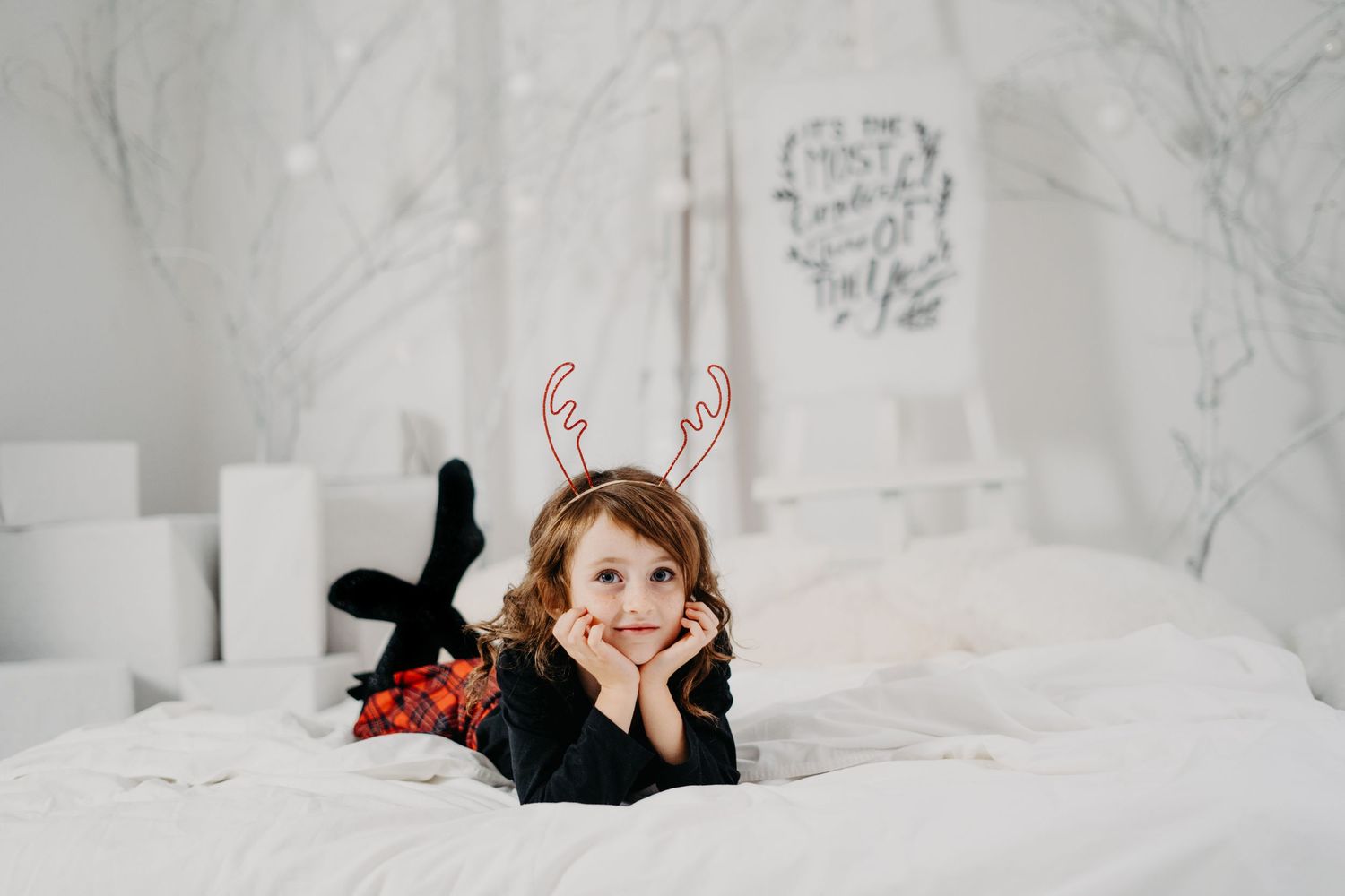 Small figure wearing reindeer antlers lies on white bedding in bright holiday themed bedroom setting.