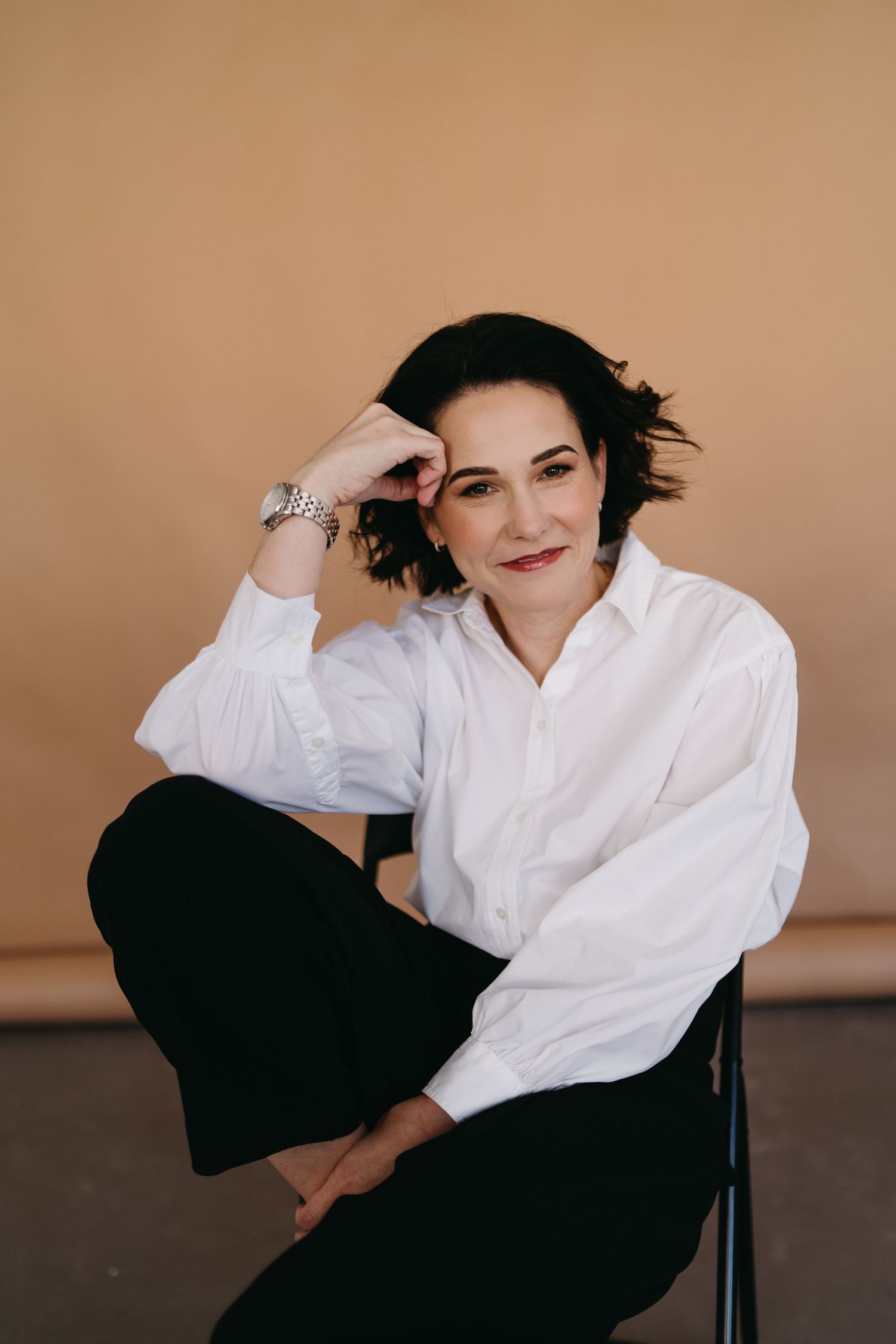 Woman in white shirt and black pants sits relaxed on chair, smiling at camera against beige background.