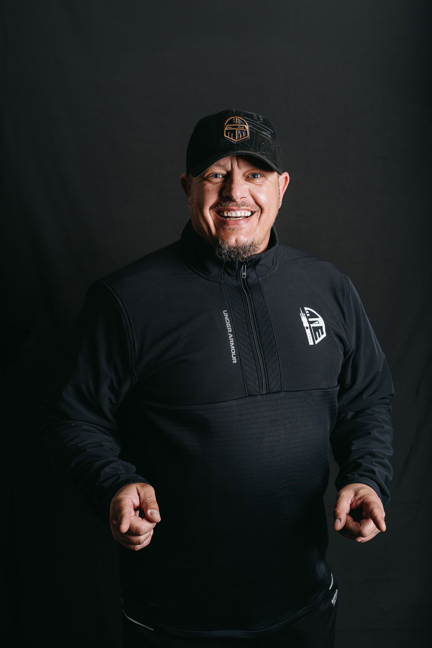 Athletic coach wearing black quarter-zip pullover and baseball cap smiles against dark background.
