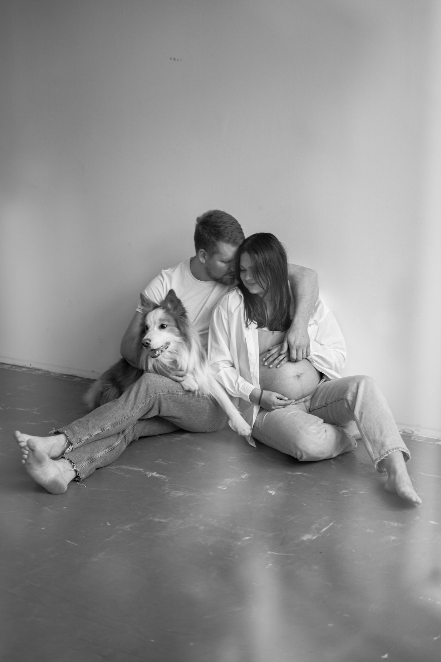 A black and white maternity photo of a couple sitting on the floor in casual attire.